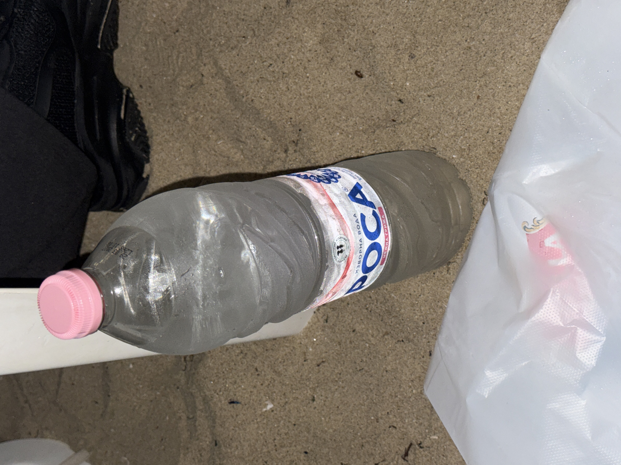 The image captures a ground-level view of a sandy environment in Varna, Bulgaria. A clear plastic water bottle with a pink screw-top cap stands centrally. The bottle contains a clear liquid and exhibits external condensation, indicating a low temperature. Its label, partially visible, displays the Cyrillic text "РОСА" (ROSA) and "ИЗВОРНА ВОДА" (SPRING WATER). In the upper right quadrant, a dark-colored athletic shoe is partially visible on the sand. A vertical white object, possibly a furniture leg or support, is present in the upper left, adjacent to the bottle. In the lower left foreground, a translucent white plastic bag lies on the sand, partially revealing a pink object inside. The ground is composed of light brown sand with scattered small debris. No individuals are depicted in the scene.