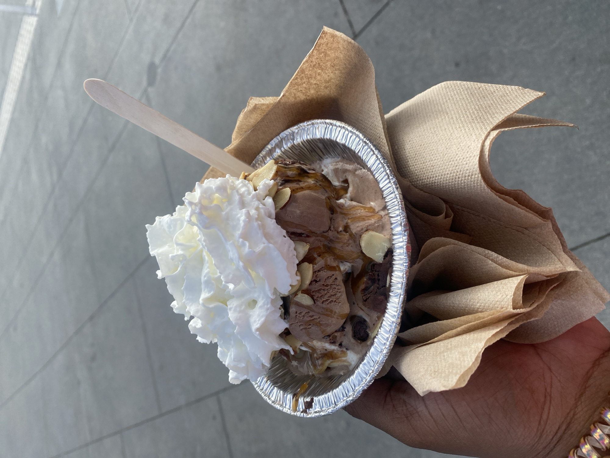 A hand holds a small, foil-lined cup of ice cream topped with whipped cream, sliced almonds, and caramel sauce, outdoors in Hermosa Beach, California. The ice cream is a mix of chocolate and vanilla flavors. A wooden spoon sticks out of the ice cream. The ground appears to be concrete pavement.