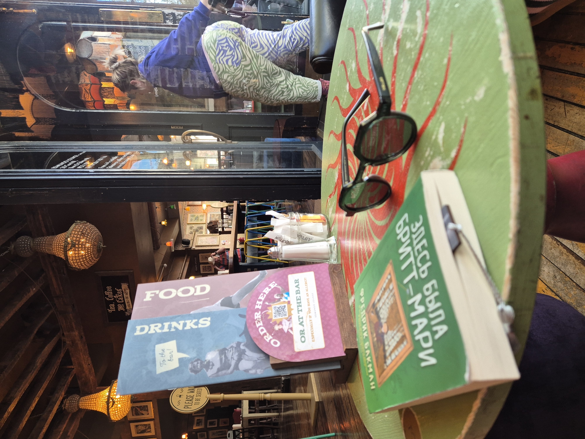 The image captures a view from within a building, likely a restaurant or cafe in West Suffolk, United Kingdom, focusing on a table with various objects. The table, painted green with a red sunburst design, holds a pair of black sunglasses, a book in Russian with the title "ЗДЕСЬ БЫЛА БРИТТ-МАРИ" ("Here was Britt-Marie") by Fredrik Backman, condiments in containers, and menus displaying "FOOD" and "DRINKS". The menus also contain the text "ORDER HERE OR AT THE BAR".
Beyond the table, a window reflects a person wearing a blue top and patterned trousers, possibly a customer. Through the window, the blurred outline of the building's exterior is visible. Inside, hanging light fixtures illuminate the interior, which features wooden beams and framed pictures on the walls. A sign indicates "Tea, Coffee & Cakes". A red seat is visible. There is a sign on the floor that says, "PLEASE WAIT TO BE SEATED".