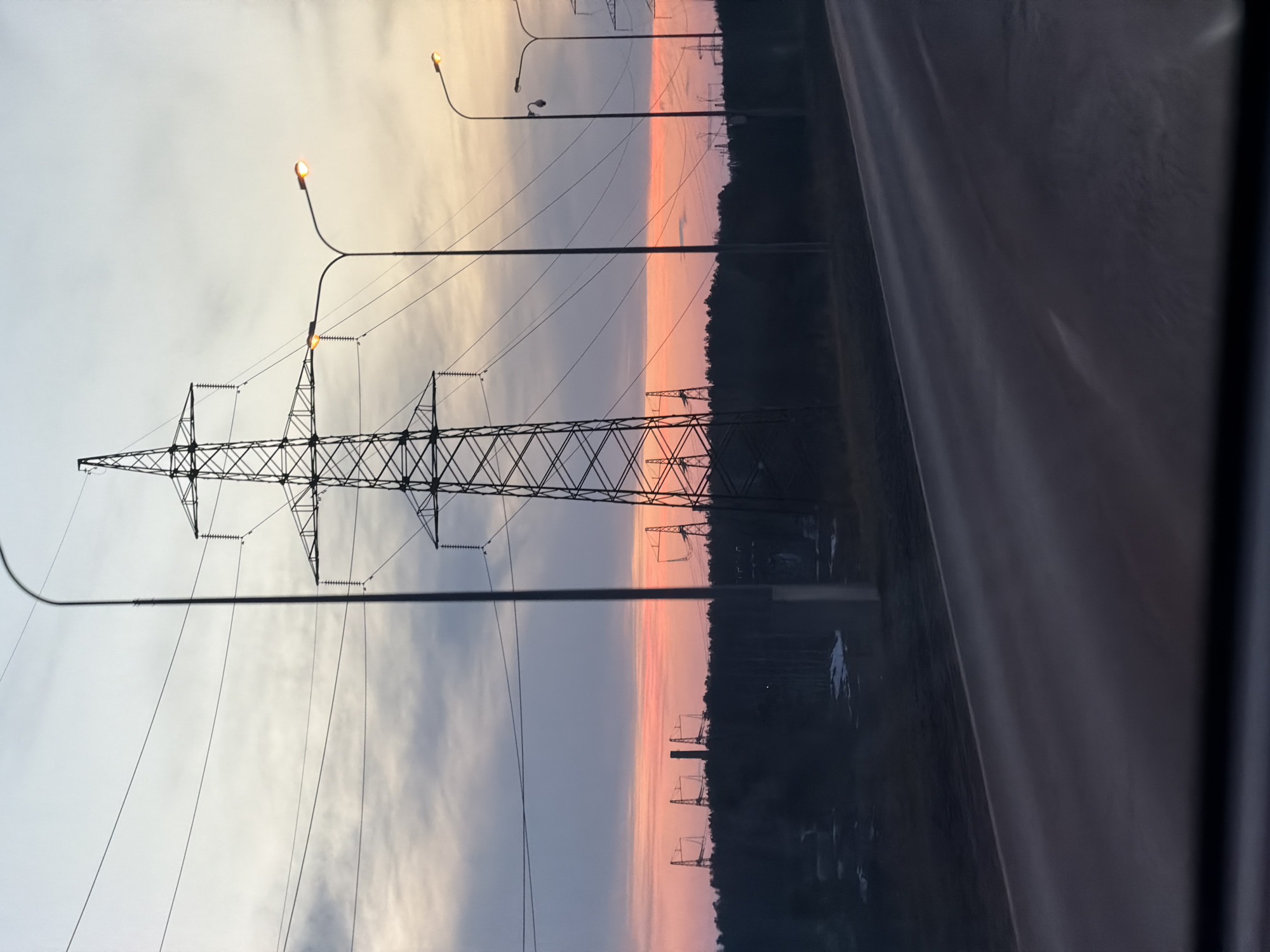 The image depicts a view of an area near the city of Chaikovsky, Russia, featuring power lines and poles against a sky gradient transitioning from light to dark, possibly during sunset or sunrise. A large electrical transmission tower is prominently visible in the foreground, with power lines stretching across the frame. Streetlights are scattered along the side of a road, some illuminated. There appears to be vegetation or a wooded area adjacent to the road. The image seems to be taken from inside a vehicle, as evidenced by the blurred interior elements in the right corner. Other transmission towers are further in the distance.