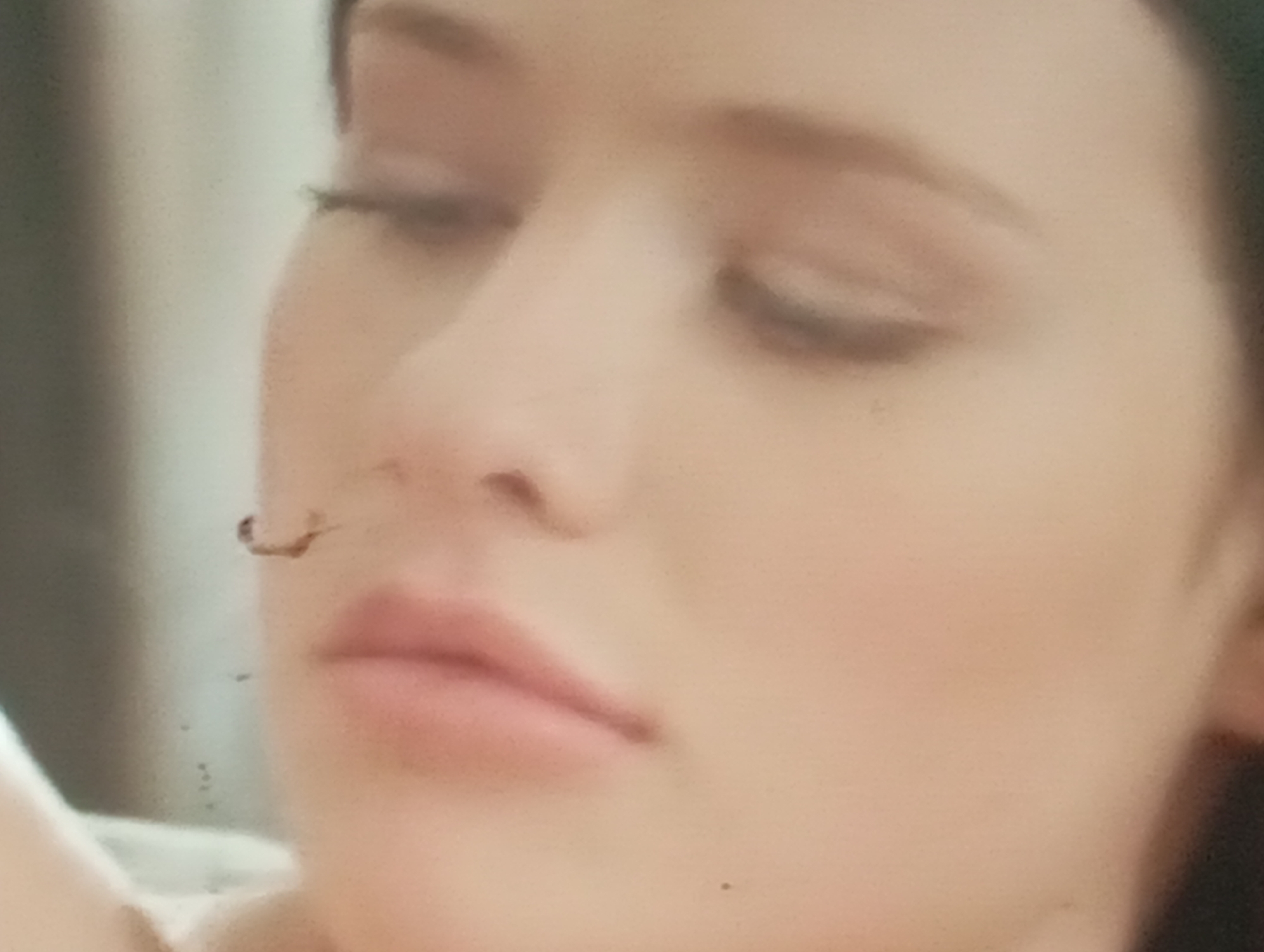 This media file is a close-up, slightly blurry photograph of a young woman's face. The subject is a person, and the setting appears to be indoors, possibly a home environment given the soft lighting and neutral background which is out of focus. The woman's eyes are partially closed, and her lips are slightly parted. Her expression could be interpreted as relaxed or pensive.
Notable details include the presence of what appears to be a small, dark, curly object near her nose, possibly a stray hair or an eyelash. There are also some small specks of dust or dirt visible on the surface of the image, particularly in the lower left quadrant. The lighting is soft and diffused, suggesting it might be natural light from a window or artificial lighting that is not harsh. There is no visible text in the image. The overall impression is intimate and personal, capturing a candid moment. Given the location context of Sofiivska Borshchahivka, Ukraine, the scene is set within this geographical area, though no specific landmarks or cultural cues are identifiable in the photograph itself. The time of day and weather are not discernible from the image.