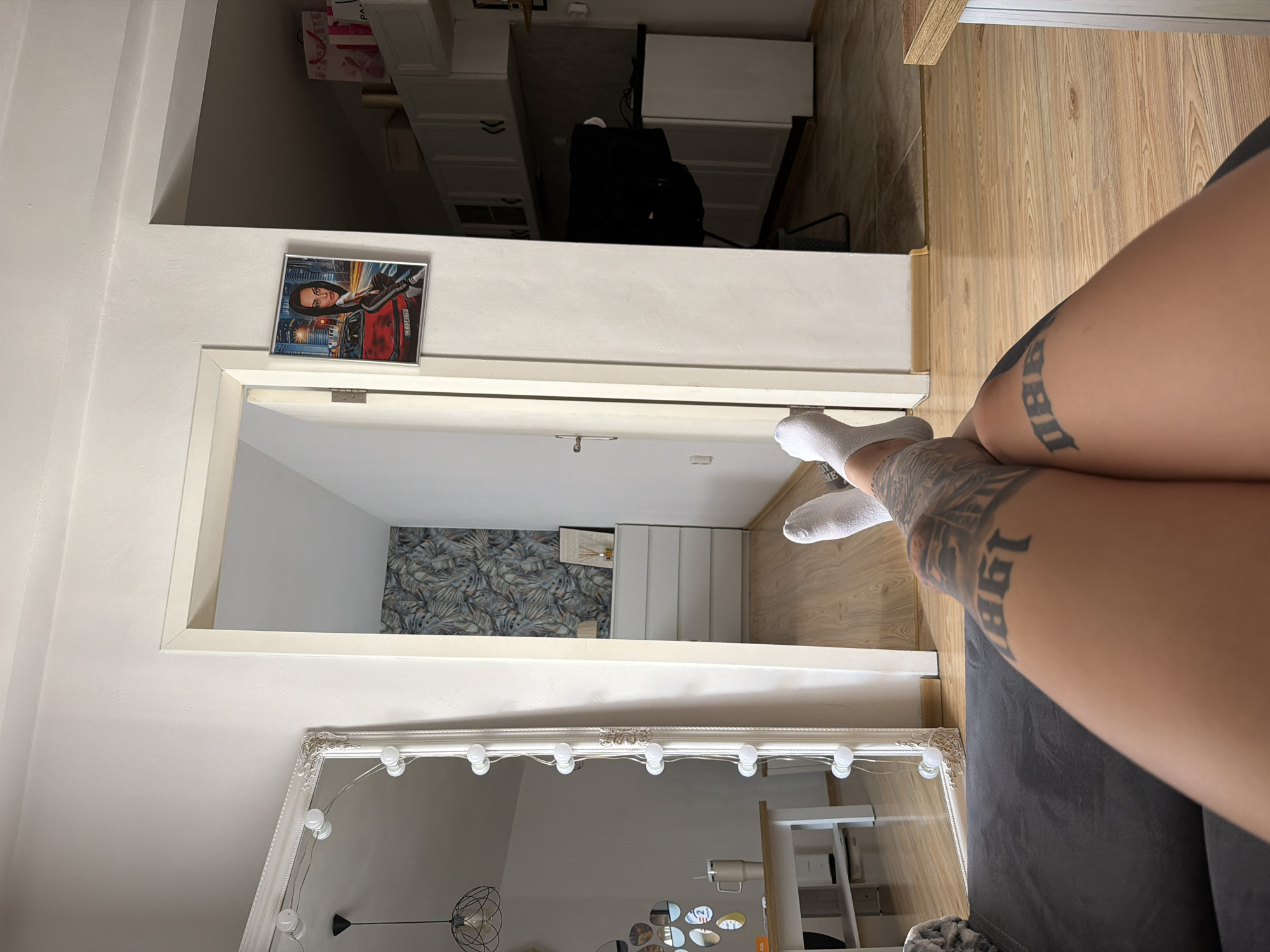 A person's legs, adorned with tattoos, are the primary subject of this image. They are wearing white socks and are positioned on a dark grey sofa. The background reveals a room with light-colored walls and wooden flooring.
In the middle ground, a doorway leads into another part of the room, where a patterned wallpaper adorns one wall. A chest of drawers is visible. Above the doorway, a framed picture is displayed, featuring a woman in a red vehicle against a backdrop of a city.
The foreground is dominated by a large, ornate mirror with lights around its frame, reflecting a ceiling lamp and other elements of the room, including a glimpse of what appears to be a white table. The overall setting suggests an indoor residential space, likely a living room or bedroom, in Plovdiv, Bulgaria. The lighting appears to be artificial, possibly from lamps and overhead lights, and there are no strong indications of the time of day or weather. The image is taken from a low angle, focusing on the person's legs and the surrounding interior. Visible text includes numbers and lettering on the tattoos.