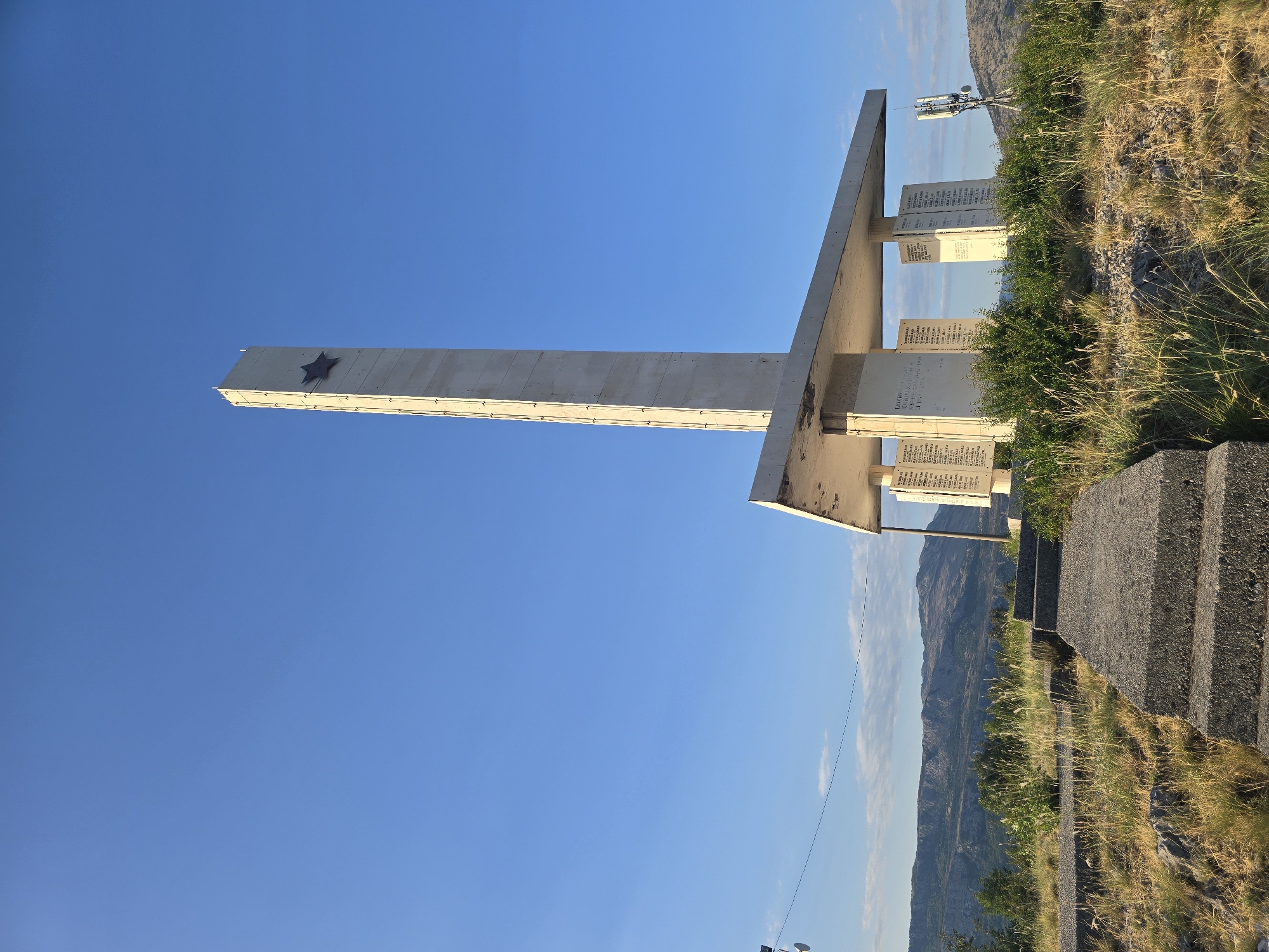 This is a full-shot, eye-level, outdoor photograph of a concrete memorial with a tall, slender obelisk topped by a red star. The memorial is situated on a grassy, rocky hilltop under a clear blue sky. Concrete steps lead up to the memorial, which has a cantilevered concrete roof supported by columns. Inscriptions are visible on panels mounted on the columns. To the right of the memorial, a cell tower is visible. In the background, rolling hills and mountains are visible under the sky.