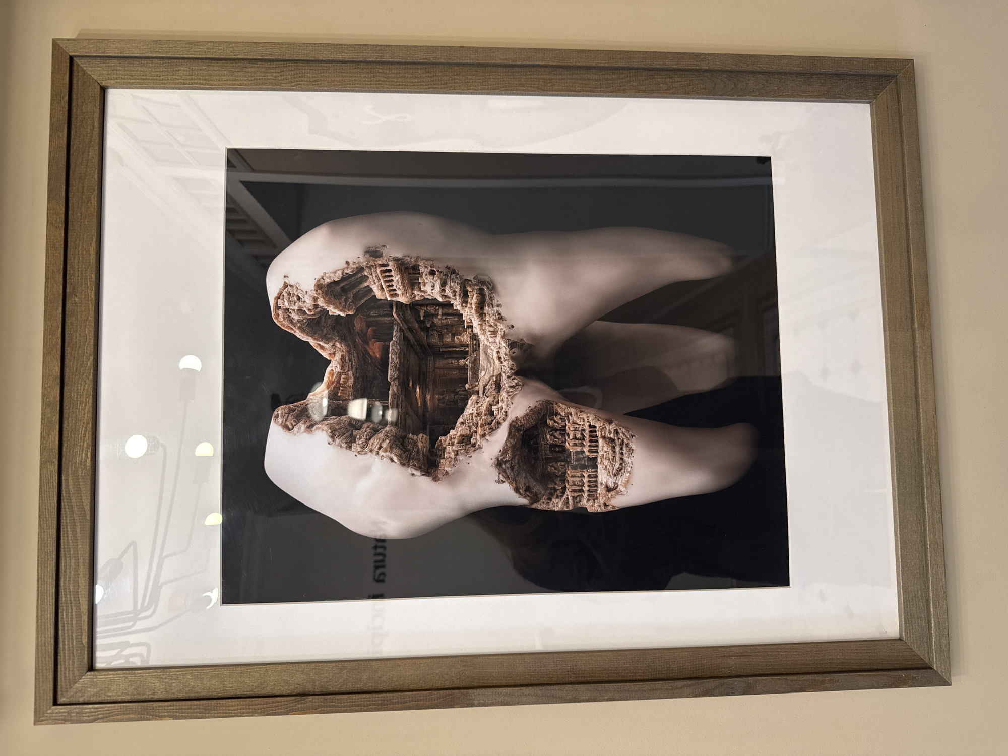 The image shows a framed artwork hanging on a beige wall. The frame is wood with a double border, the inner border being white. The artwork within depicts a molar-shaped object with an interior that appears to be a complex architectural structure. The interior has brown tones suggesting stone or wood. Reflected in the glass covering the artwork is a partial view of a light fixture with multiple arms and lights. Text in the Thai language is visible at the bottom of the artwork. There is no indication of the image being taken in the city of Izhevsk, Russia.