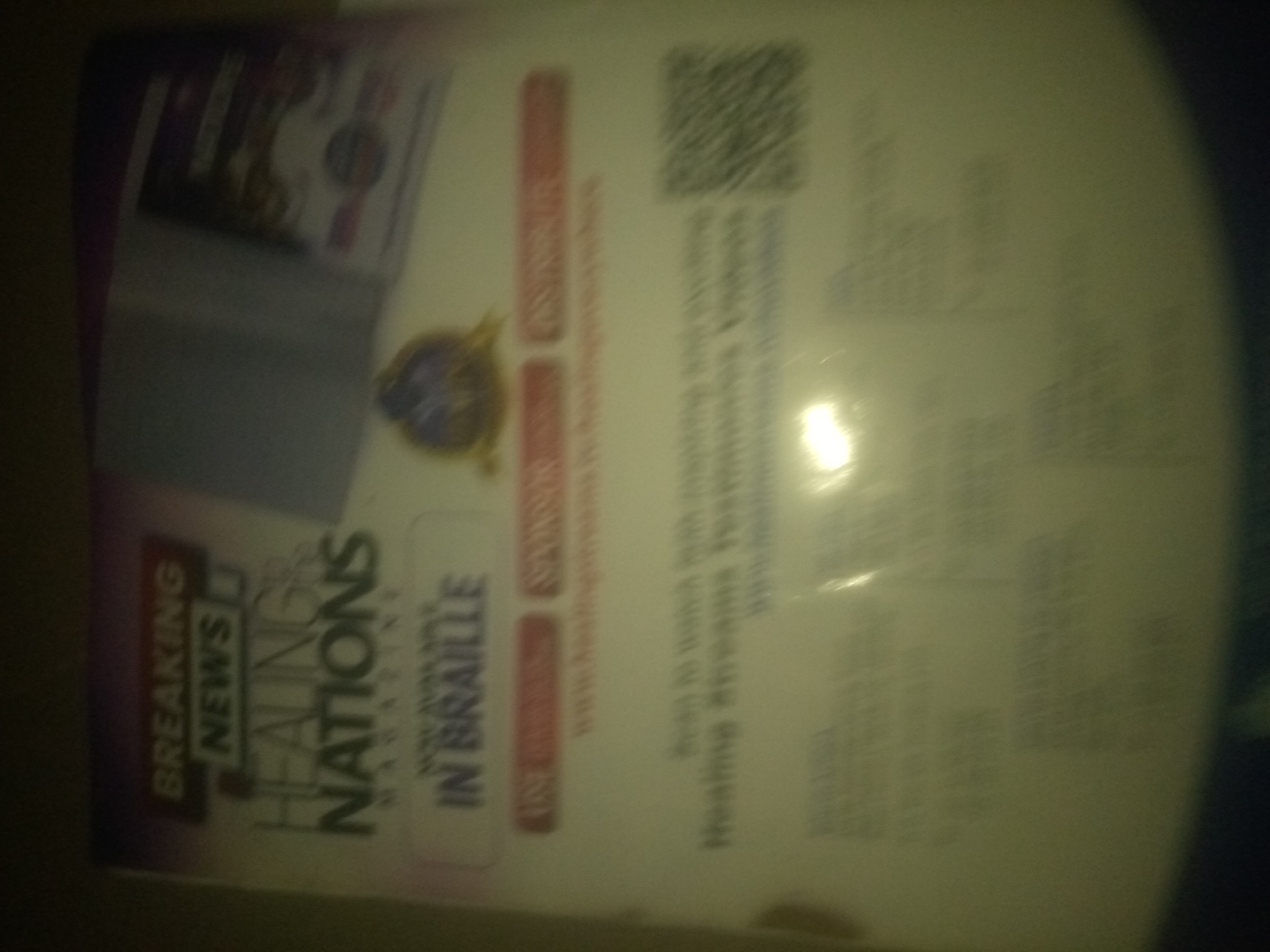 The image shows a portion of a magazine or publication. The text "BREAKING NEWS" and "HEALING NATIONS" are visible. The publication appears to be a magazine titled "HEALING NATIONS MAGAZINE" and also states "IN BRAILLE". A QR code is present in the upper right quadrant of the image. The background is a plain white surface, possibly a table or wall. The image is taken at an angle and is slightly blurry, making it difficult to discern finer details. There are no people or specific environmental details visible beyond the product itself.