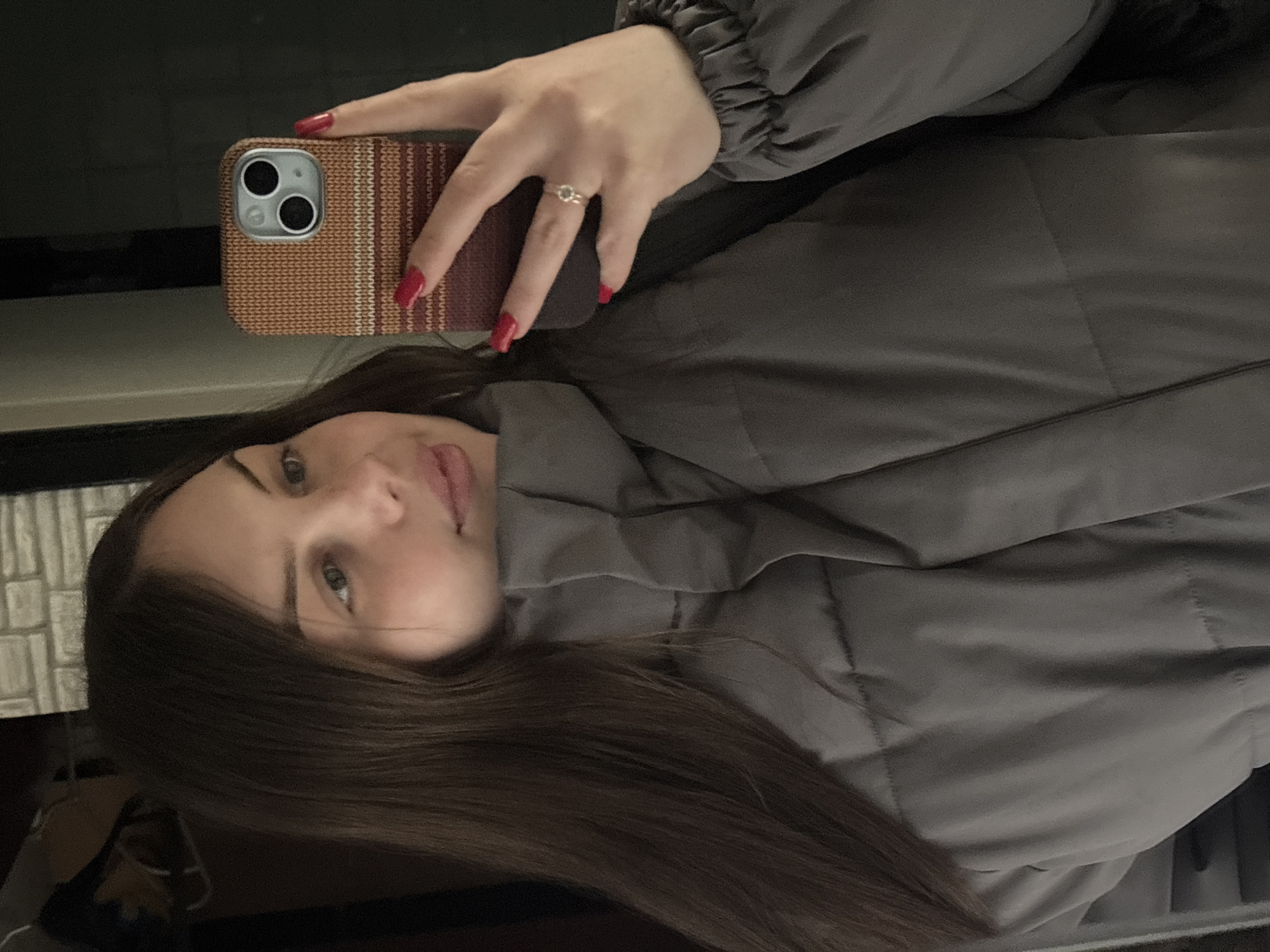 A woman is taking a selfie in Новосельское сельское поселение, Ukraine. She is wearing a gray puffer jacket and holding a cell phone with a patterned case. She has brown hair and red nail polish. A ring is visible on her finger. The background is dark and slightly reflective.