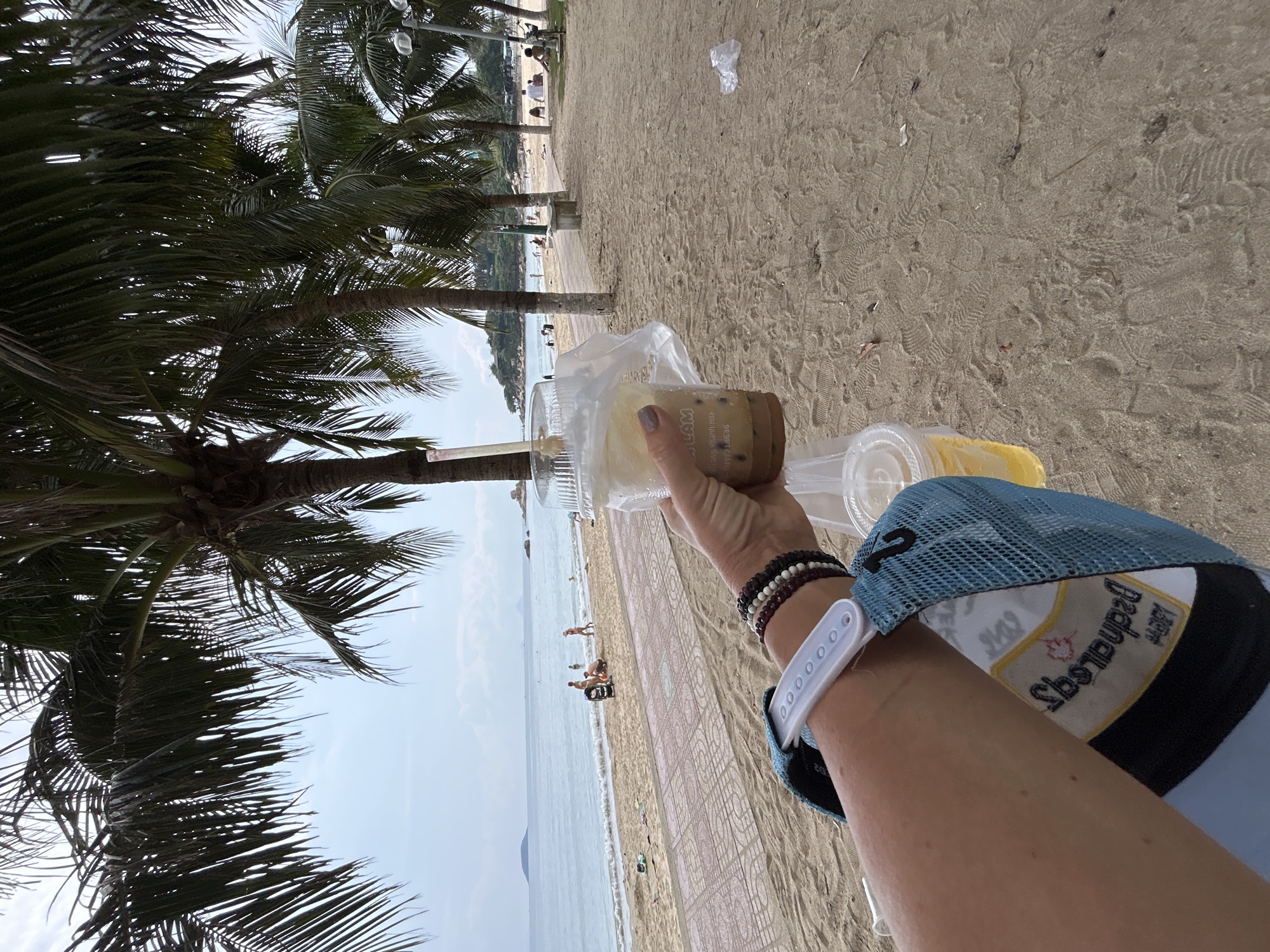 View from a beach in Bắc Nha Trang, Vietnam. The image appears to be a first-person view of an arm holding a beverage on a sunny day. The beach is visible in the background, along with palm trees, the ocean, and the sky. Other people are present on the beach. The arm has a cap.