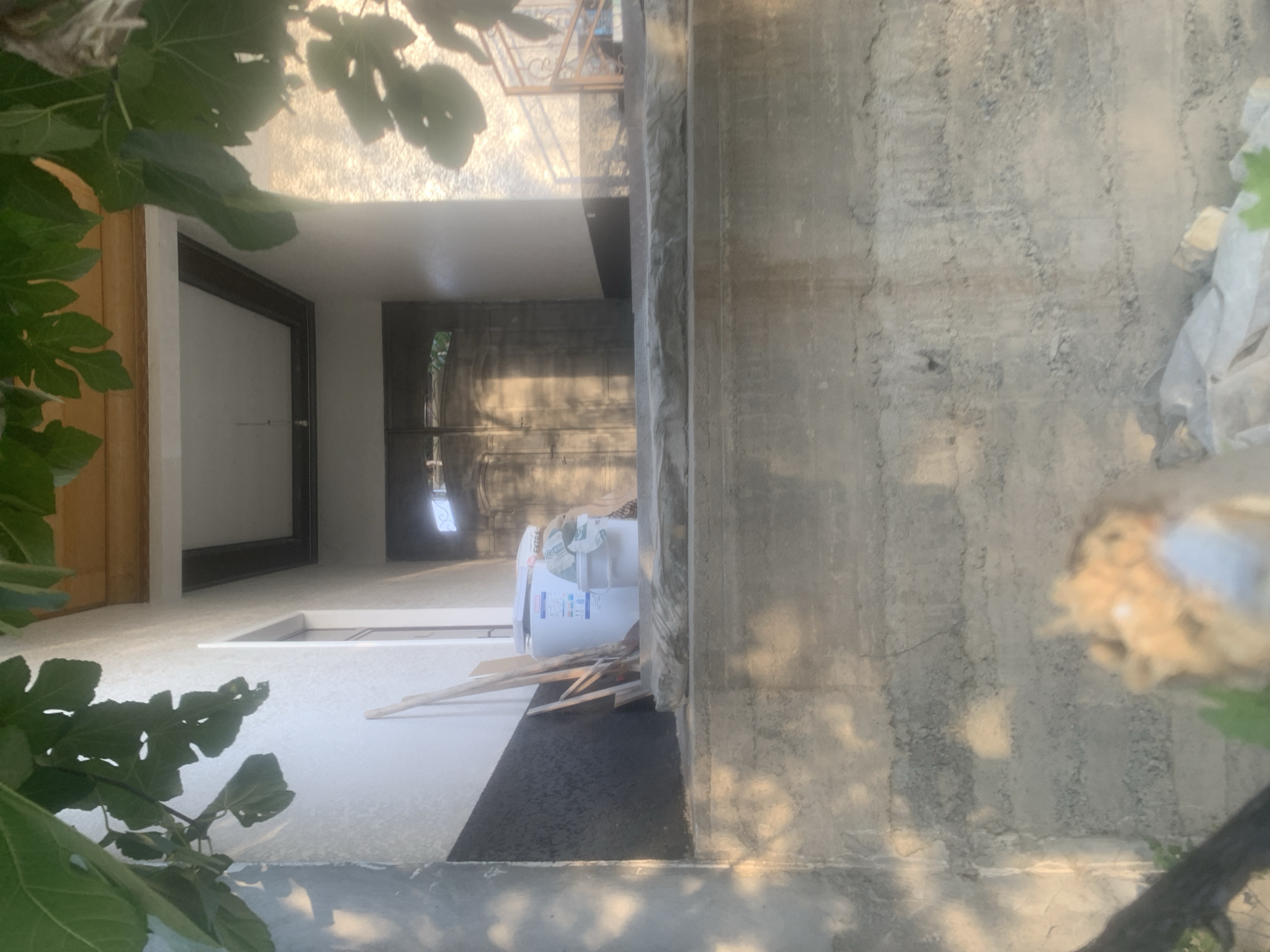 The image shows an outdoor scene, likely in a residential area given the presence of a house entrance. In the foreground, a textured concrete wall is visible, with dappled sunlight creating patterns of light and shadow. Above this wall, a raised concrete platform leads to the entrance of a building. To the left of the platform, against a white wall with a black lower section, are several wooden planks leaning upright, a white bucket, and a white cylindrical object, possibly a water heater, with some labels. Beyond this, a walkway leads to a large, dark double door with ornate metal detailing. The entrance is recessed and covered by a roof supported by columns. To the right of the entrance, a decorative metal railing is visible, suggesting a staircase or elevated walkway. Green foliage, likely from a tree, hangs over the scene, casting shadows and partially obscuring the view. The overall impression is one of a humble dwelling with some construction or renovation materials present. The lighting suggests it is either early morning or late afternoon.