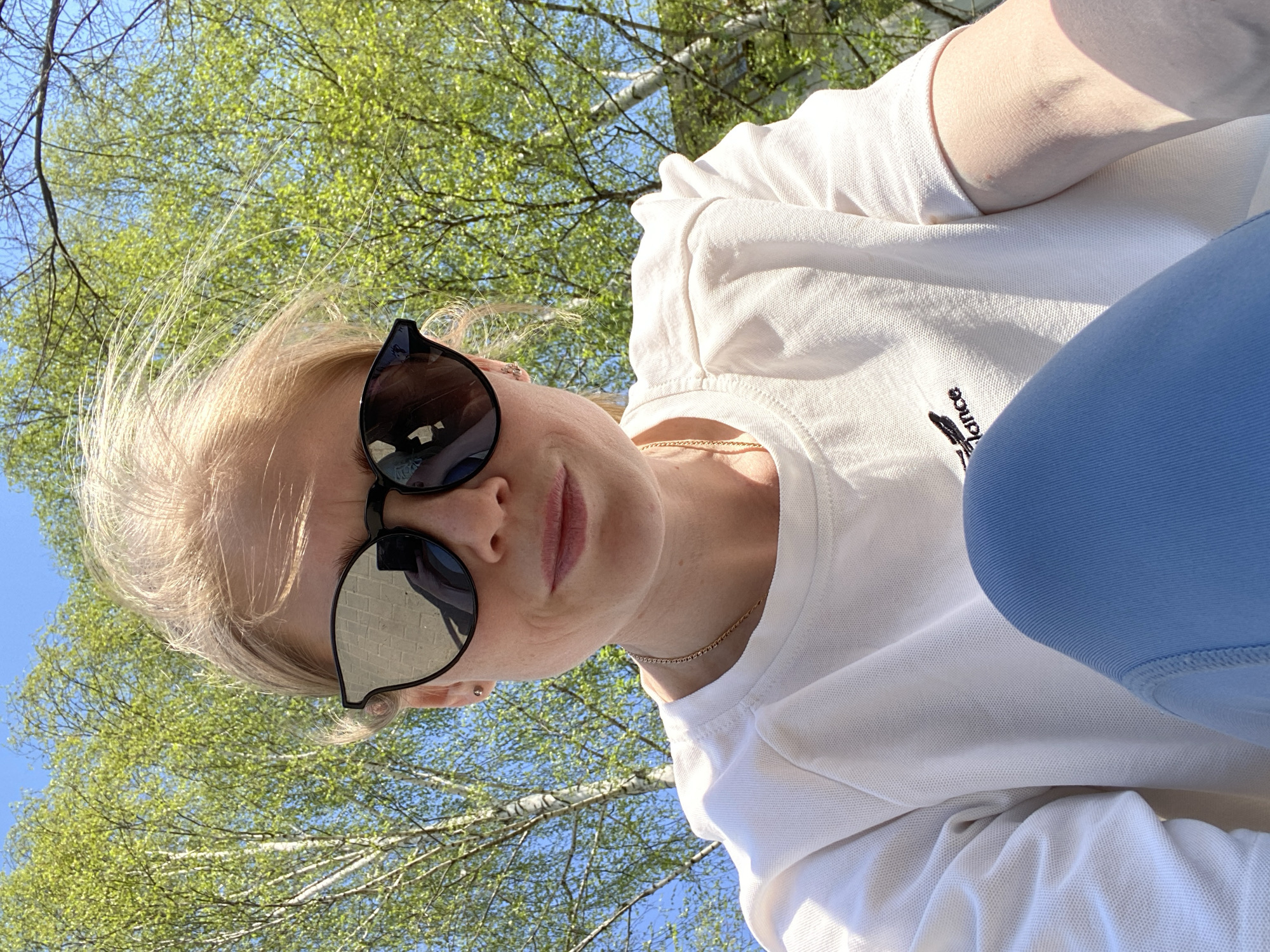 A fair-skinned, blond-haired woman wearing sunglasses, a white t-shirt, and blue pants poses in an upward-angled selfie outdoors. A thin gold chain is visible around her neck. The background features a blue sky interspersed with green tree leaves and white birch tree branches. The location cannot be determined as Chaikovsky, Russia from the image.