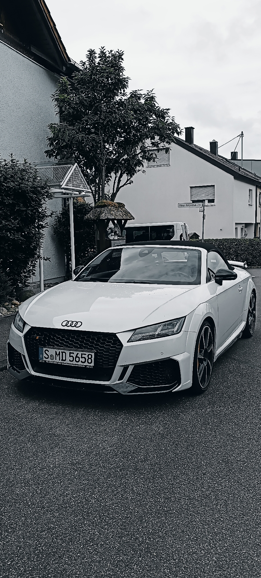 A white Audi TT convertible is parked on a wet asphalt street in front of a residential building. The car has its top down and the license plate reads "S MD 5658". To the left of the car is a tree and a tall pole with a triangular structure at the top. Behind the car, a white building with a sloped roof and dark-trimmed windows is visible. A street sign reads "Obmar-Morgenhaler-Str.". The overall scene is outdoors on a cloudy day. The location is identified as Waiblingen, Germany.