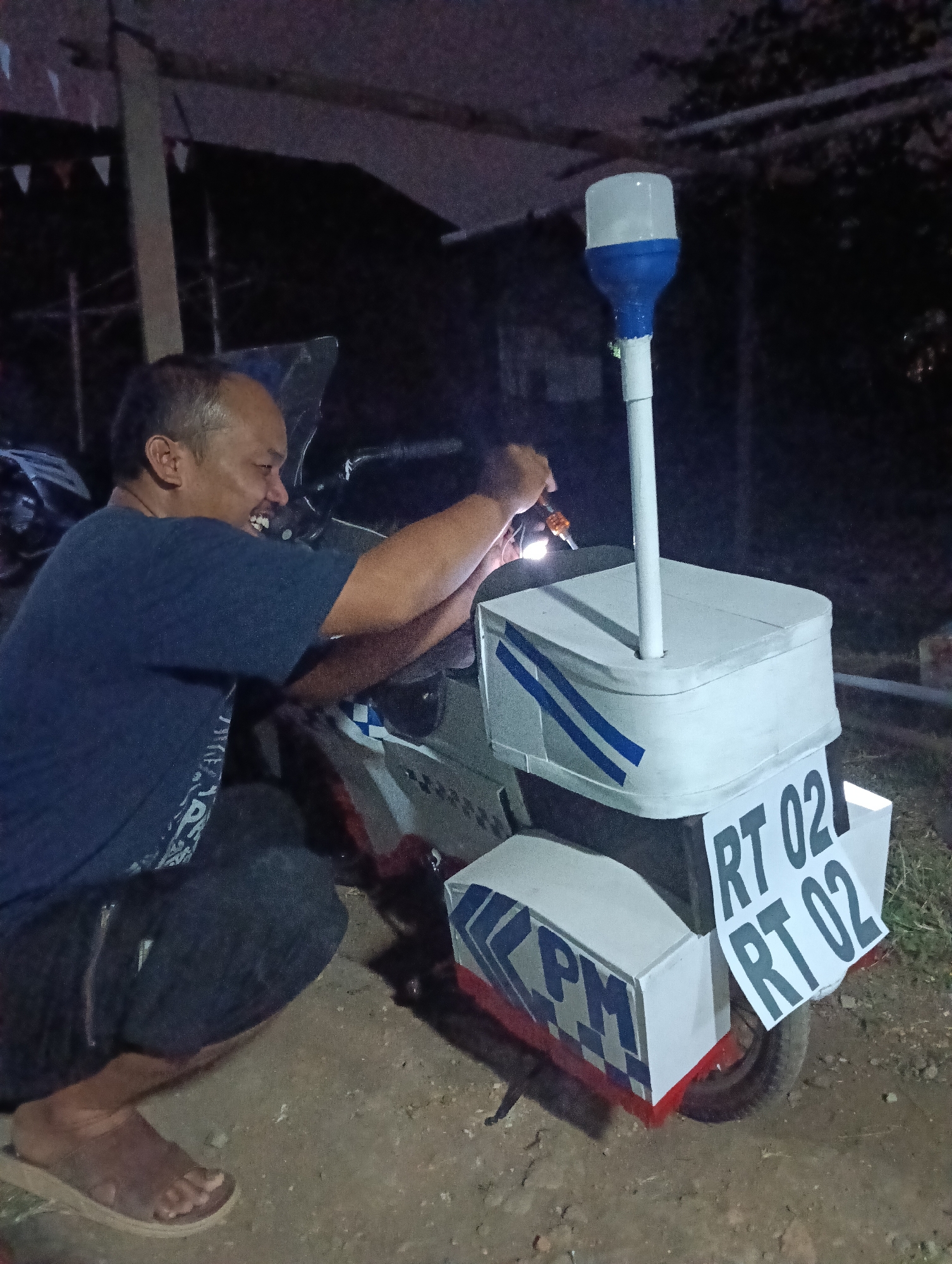 An Indonesian man is modifying a white scooter in an outdoor, dimly lit environment. The scooter has been fitted with additional compartments and a blue and white light pole on top. The man, wearing a dark blue t-shirt and dark pants, is crouched down and appears to be working on electrical components of the scooter, holding a tool that emits light. The scooter has the labels "RT 02 RT 02" and "KPM" on its side compartments. The ground is rough and dirt-like. The background shows some vegetation and wooden structures. The presence of "RT" and "KPM" suggests this is likely related to community organization or public services within Tasikmalaya, Indonesia, as these are common designations for neighborhood and village administrative units.