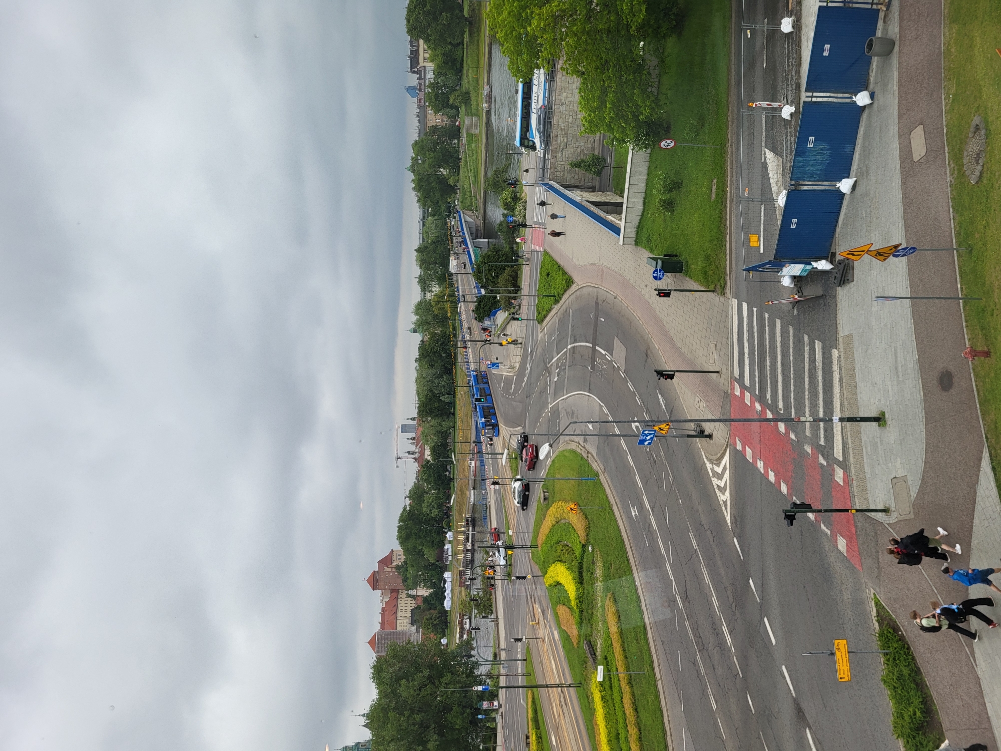 This high-angle, wide shot captures a panoramic view of an urban landscape in Kraków, Poland, on an overcast day. The scene focuses on a complex road intersection, the Vistula River, and the Wawel Royal Castle in the background.
In the upper-left and center-background, the Wawel Royal Castle, characterized by its reddish-brown tiled roofs and fortified walls, stands prominently on a hill. A green-domed spire of a church or building is visible to the far left of the castle. Beyond the castle, distant city buildings and a construction crane are discernible against the cloudy, light grey sky.
The Vistula River flows through the midground, with several boats or barges docked along its far bank. Two bridges span the river: the Most Dębnicki bridge, identifiable by traffic, and another bridge further to the right. A blue tram is visible on the left side of the main bridge, and a light blue "Trans-Usługi" bus is positioned on the bridge to the right.
The foreground is dominated by a multi-lane asphalt road network featuring white lane markings and a prominent red-painted pedestrian crosswalk with white stripes. A large, landscaped traffic island in the center of the intersection contains ornamental plantings in yellow and green. Multiple traffic lights, streetlights, and various European road signs are present, including a yellow warning sign for cyclists pointing towards "Most Dębnicki / Modliborzycka / Wisła-Odra" and a "40 km/h" speed limit sign. Several vehicles, including white, red, and dark-colored cars, are visible on the roads.
On the sidewalks, composed of grey pavers, pedestrians are observed walking. In the bottom-right corner, blue temporary construction barriers, secured with white sandbags, line a section of the sidewalk, with orange cones and warning signs nearby. A red fire hydrant is visible in the lower-left corner. The environment is rich with lush green trees and grassy areas, particularly along the riverbanks and adjacent to the roads.