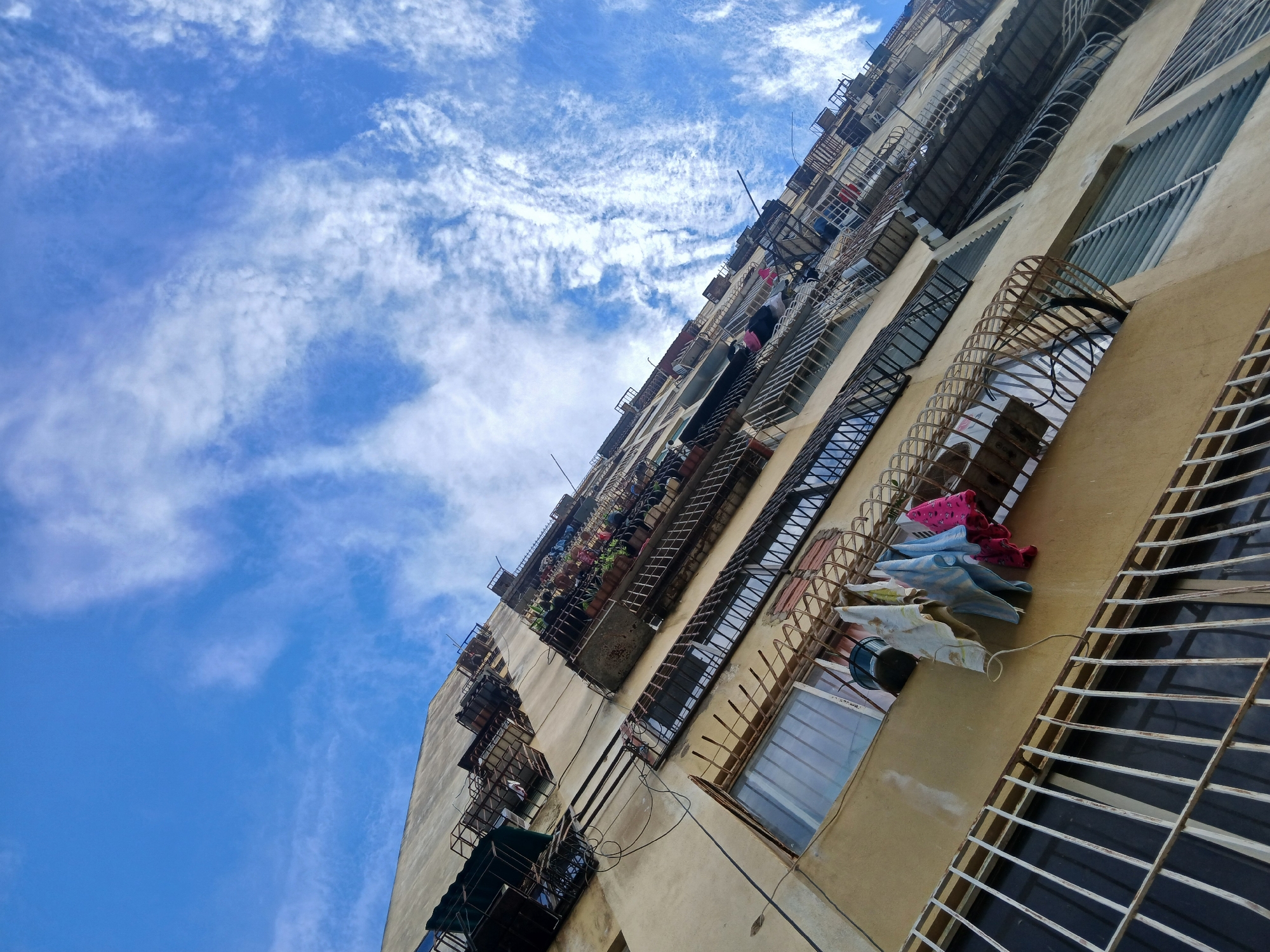 This image depicts a multi-story, light-colored apartment building in Ciudad Guayana, Venezuela, captured from a low, upward-looking perspective against a bright blue sky with scattered white clouds. The building's facade is characterized by numerous windows and balconies, many of which are fitted with rusted, dark metal security bars or grates. On a lower floor, five small items of laundry—including white, blue, and pink garments—are visible hanging to dry from a rusted metal grate. Higher up, multiple balconies feature potted plants. Exposed electrical wiring and conduits are present on the building's exterior. No individuals are visible. The overall impression is one of urban residential architecture under daylight conditions.