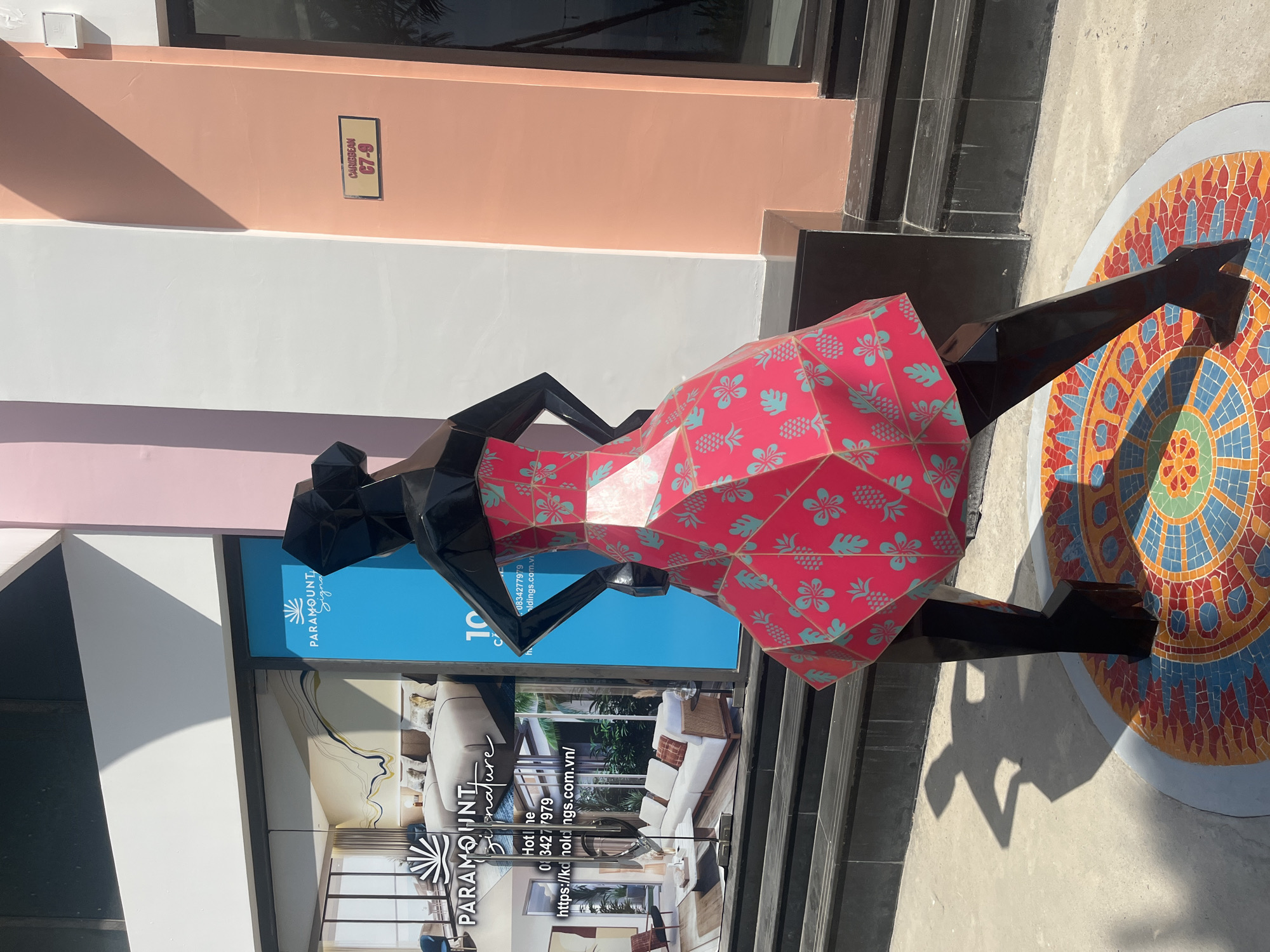 The image depicts an outdoor scene in Nha Trang, Vietnam, featuring a striking geometric sculpture of a woman in a vibrant pink dress adorned with a turquoise pineapple and floral pattern. The sculpture stands on a colorful mosaic circular base. In the background, a modern building features large glass windows displaying interior renderings of hotel rooms, with signage for "PARAMOUNT Signature". Adjacent to this, a bright blue panel displays the text "PARAMOUNT Signature" with a logo, and further to the right, a peach-colored wall has a sign that reads "CARIBBEAN C7-9". The sun casts a clear shadow of the sculpture on the concrete ground. The environment suggests a commercial or hospitality area, likely a promenade or entrance to a resort or hotel.