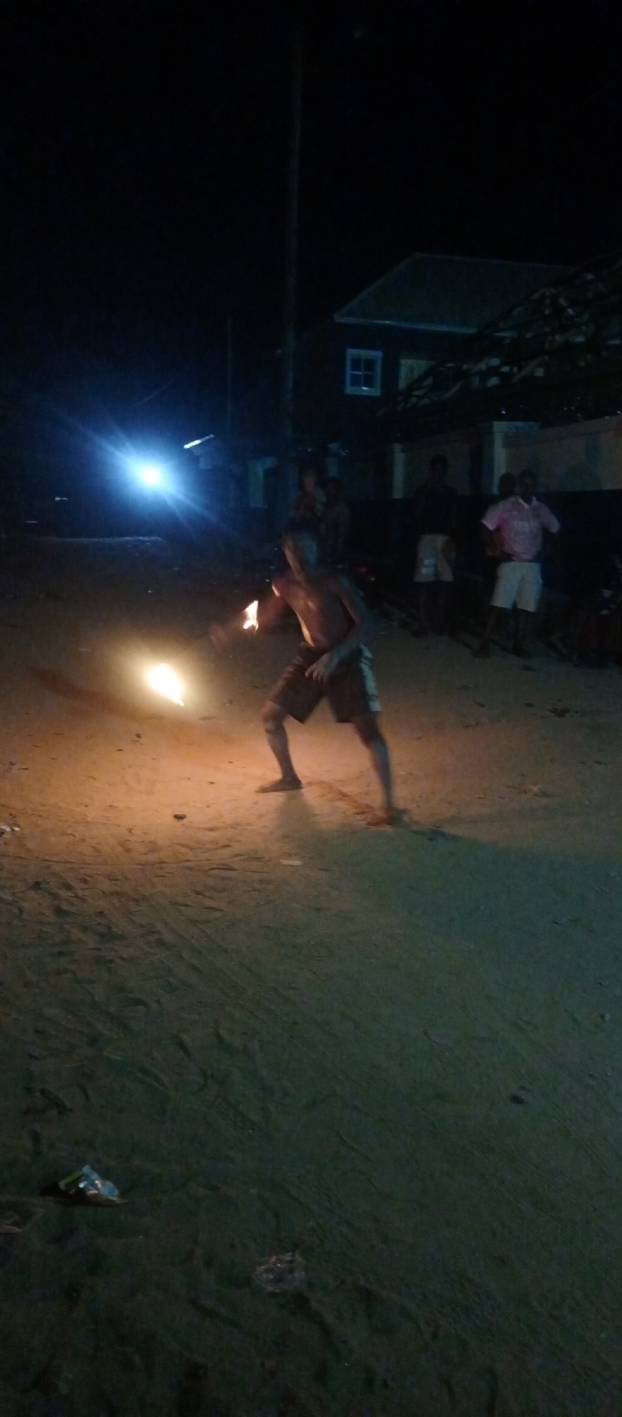 The image depicts a nighttime scene in Keffi, Nigeria, focusing on a fire performer in the foreground. A shirtless individual, likely a young man or boy, wearing dark shorts, is actively engaged in fire dancing or performing. He holds a flaming object in his right hand, and another glowing fire source is visible near his left foot, casting a bright orange light on the sandy or unpaved ground around him. His stance suggests dynamic movement or a performance in progress.
In the mid-ground, several other people are visible, dimly lit, appearing to be spectators of the fire performance. One individual on the right wears a pink shirt and white shorts. A bright, artificial light source, possibly a vehicle headlight or street lamp, shines intensely from the mid-ground left, creating a prominent lens flare.
The background features a dark building with a white-framed window and a dark roof, along with what appears to be a utility pole. The ground is rough and unpaved, with some scattered litter (plastic waste) visible in the immediate foreground. The overall setting is dark, illuminated primarily by the fire and other artificial lights, indicating it is late evening or night. No text is visible in the image.