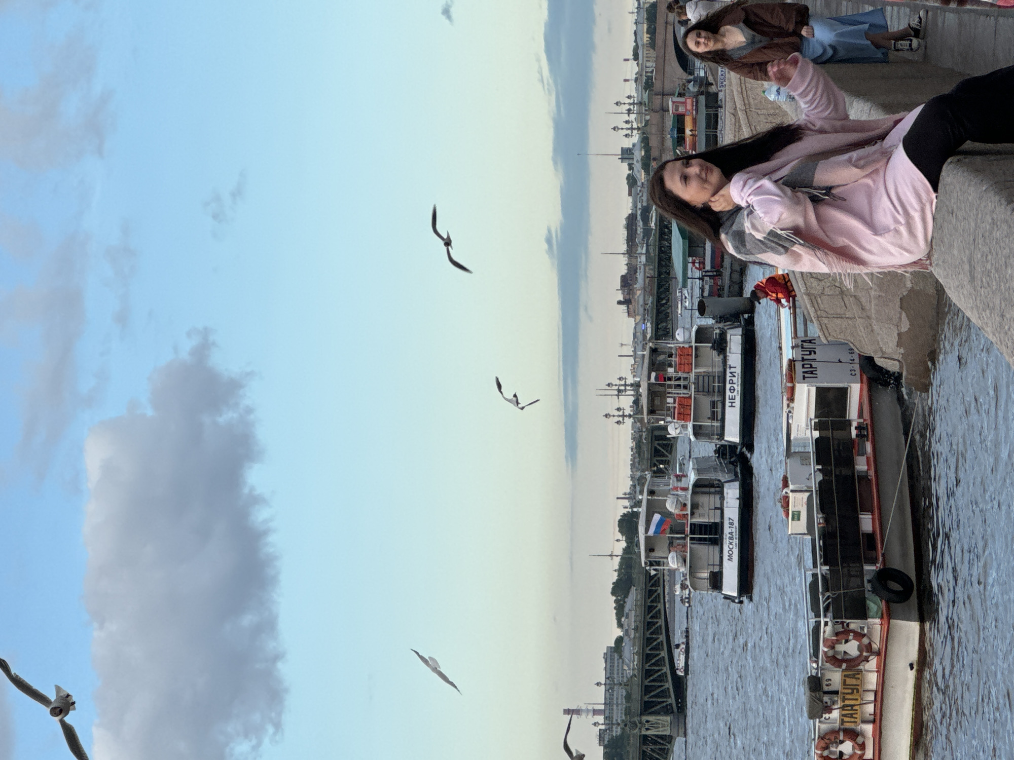 The image shows a waterfront scene in Saint Petersburg, Russia, likely during the evening as indicated by the soft, diffused light and the sky transitioning from blue to a pale yellow. Several people are visible, some seated or lounging on a stone embankment in the foreground, while others are on boats in the water. One young woman in a pink jacket and scarf is the most prominent subject, looking towards the camera with a relaxed expression. Another woman is seated behind her, wearing a brown jacket and blue pants.
Several boats, likely passenger vessels, are docked along the embankment. One boat has "ТАРТУГА" and "C3-114-68" visible on its side. Another boat is marked "МОСКВА-187" and "НЕФРИТ". Russian flags are displayed on some of the boats. Seagulls are flying in the sky, adding to the atmosphere of a coastal city. The overall mood is calm and leisurely, suggesting a pleasant evening spent by the water. The architecture in the background hints at a European city, consistent with the Saint Petersburg location. The water appears calm, reflecting the sky.