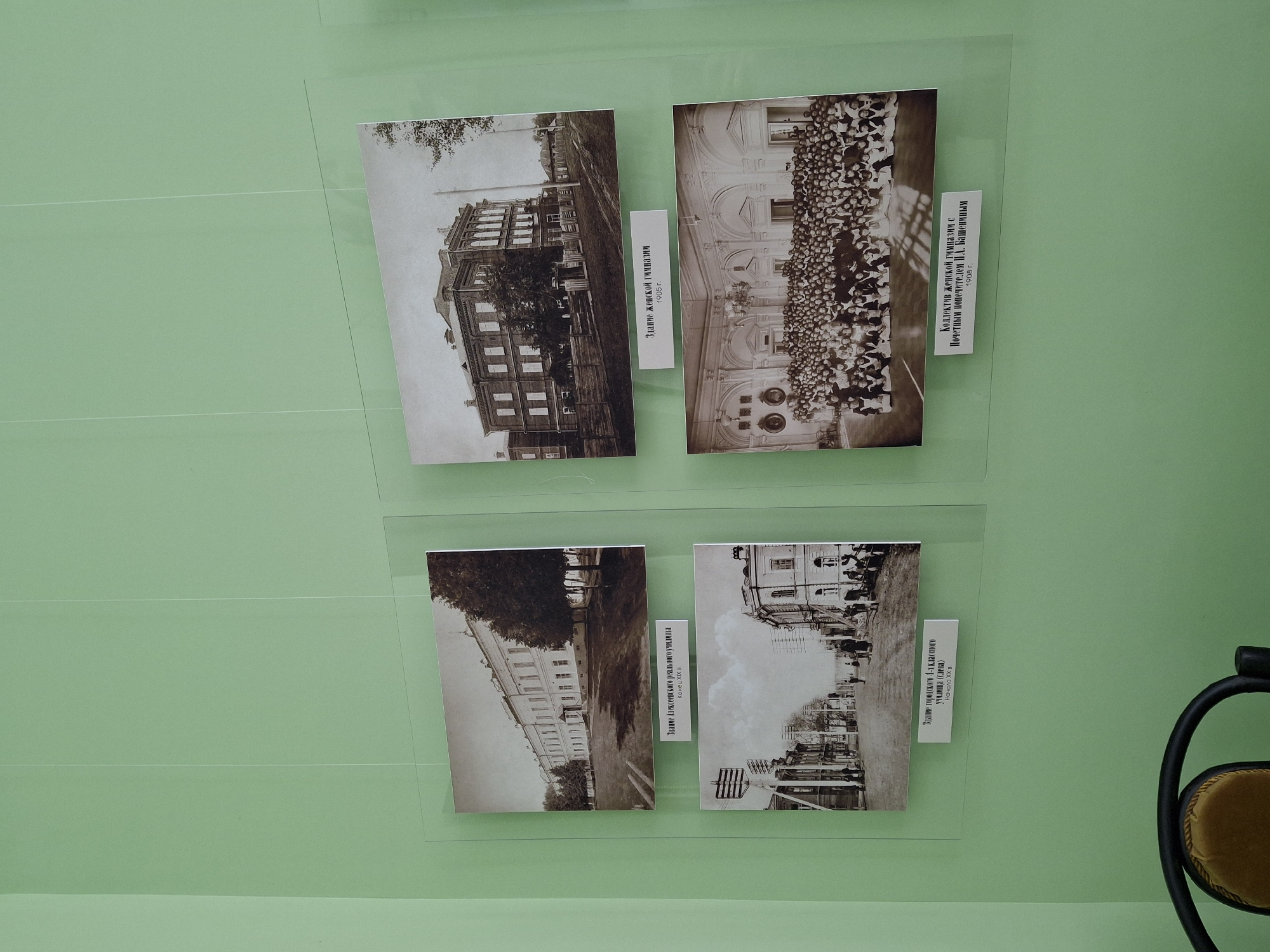 This media file displays an exhibit on a light green wall, featuring four historical black and white or sepia-toned photographs, likely from Borok, Russia. Each photo is under a transparent protective layer and accompanied by a caption.
The top-left photograph shows a multi-story wooden building with many windows, possibly a school, with trees visible around it. Its caption below reads: "Здание женской гимназии 1905 г." (Building of the Women's Gymnasium 1905).
The top-right photograph depicts a large group of people, predominantly young women, arranged in rows posing outdoors in front of a grand, ornate building. The caption to its right states: "Коллектив женской гимназии с Почетным попечителем П.Л. Башениным 1908 г." (Collective of the Women's Gymnasium with Honorary Trustee P.L. Bashenin 1908).
The bottom-left photograph features a long, multi-story building with a light-colored facade and numerous windows, surrounded by trees. The caption below it says: "Здание Алексеевского реального училища Конец ХІХ в" (Building of the Alekseevsky Real School, End of 19th century).
The bottom-right photograph shows a street scene from an early 20th-century town. Buildings line the street, with utility poles and wires visible, and a few individuals can be seen walking. The caption to its right reads: "Здание городского 4-х классного училища (слева) Начало ХХ в." (Building of the city 4-class school (left), Early 20th century), indicating a specific building within the street view.
The overall presentation suggests a historical exhibition, showcasing architectural and social aspects of Borok from the late 19th to early 20th centuries. A black chair with a yellow seat is partially visible on the far right.