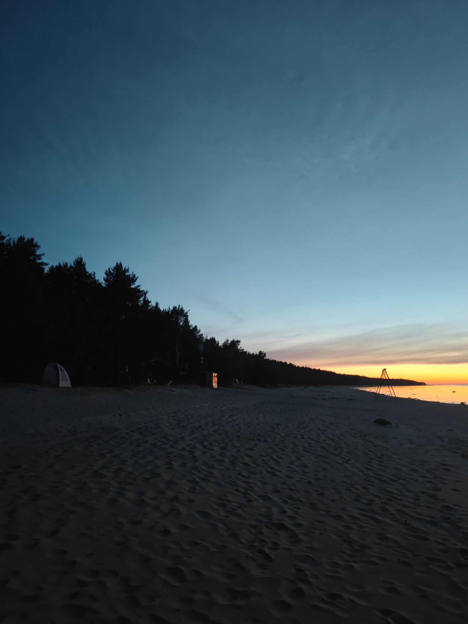 This image depicts a serene beach scene at dusk in Motornoe, Russia. The sky transitions from a deep blue to a soft gradient of orange and yellow near the horizon, indicating the setting sun. A dense line of dark trees forms a silhouette along the coastline, contrasting with the lighter hues of the sky and water.
On the sandy beach, a small, light-colored tent is pitched on the left side of the frame, partially obscured by the treeline. Further along the beach, towards the right, a tripod-like structure made of metal stands against the sunset. The sand itself is textured with ripples and footprints, suggesting recent activity. A faint orange glow emanates from what appears to be an opening within the trees, possibly a structure or a campfire. The calm water of the sea is visible on the right, reflecting the warm colors of the sky.