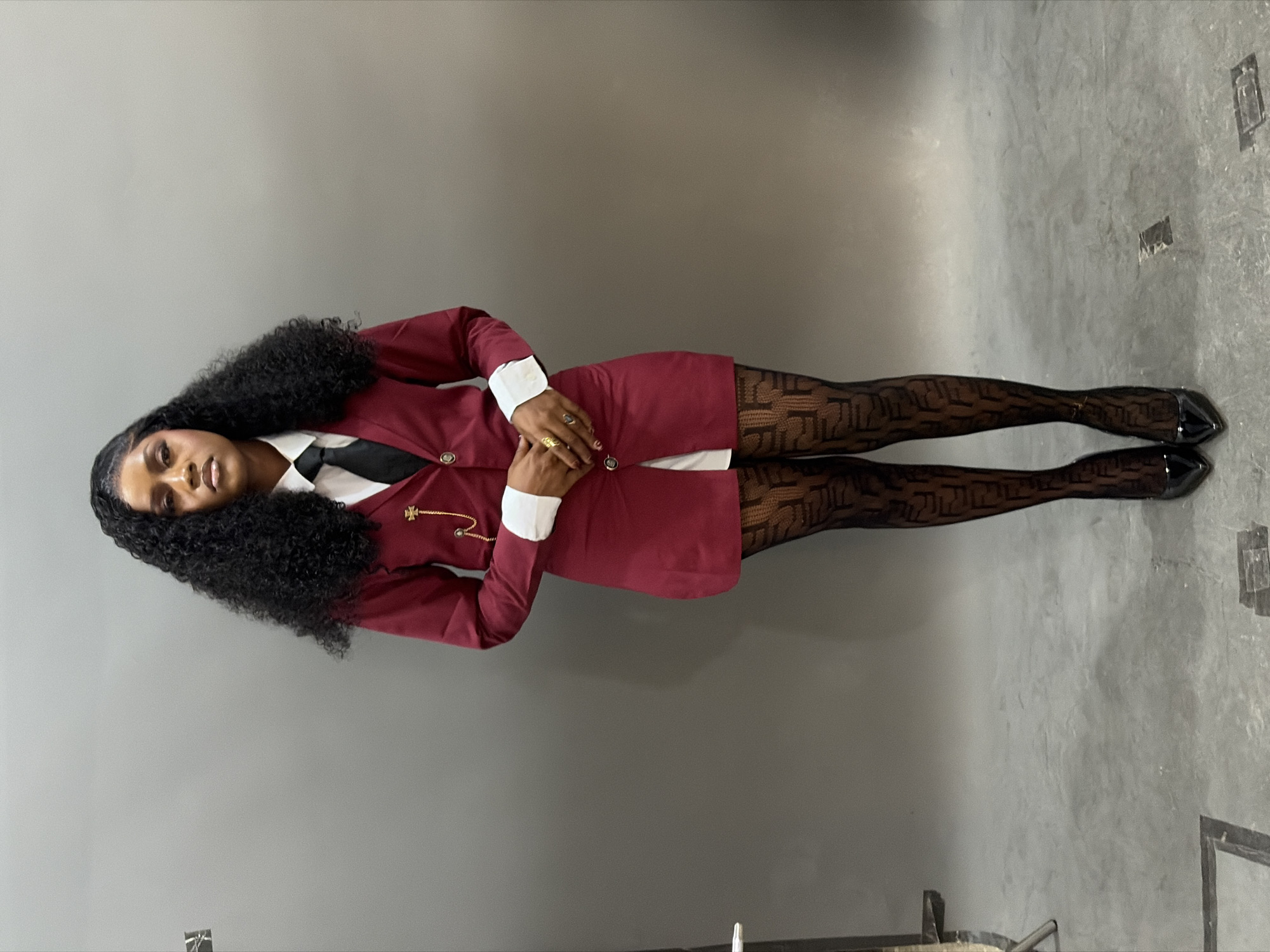 A dark-skinned individual, appearing to be a woman, is depicted in a full-body portrait, centrally positioned against a uniform gray studio background in the city of Gura Top, Nigeria. She has voluminous, dark, tightly curled hair styled around her face and shoulders, with a clear part on her left side. Her facial features include defined eyebrows, noticeable eye makeup, and light-colored lipstick; her gaze is directed straight at the camera with a neutral expression. She is dressed in a white collared shirt with visible white cuffs, a black necktie, and a short-length maroon/burgundy blazer-style dress or tunic with dark buttons. A decorative gold-colored pin resembling a Maltese cross is attached to the left lapel of the blazer, from which a delicate gold-colored chain extends. Her hands are clasped at waist level, displaying multiple gold-colored rings on her fingers and dark-painted fingernails. She wears sheer black tights or stockings adorned with a repeating pattern, specifically the 'FF' logo. Her footwear consists of black patent leather high heels with pointed toes. The floor is a continuation of the gray studio surface, exhibiting some minor scuffs and linear markings. On the far left, a portion of a metallic studio light stand or similar equipment is visible, along with a metallic door handle or latch. The scene is evenly lit, indicative of a controlled indoor photographic setting.