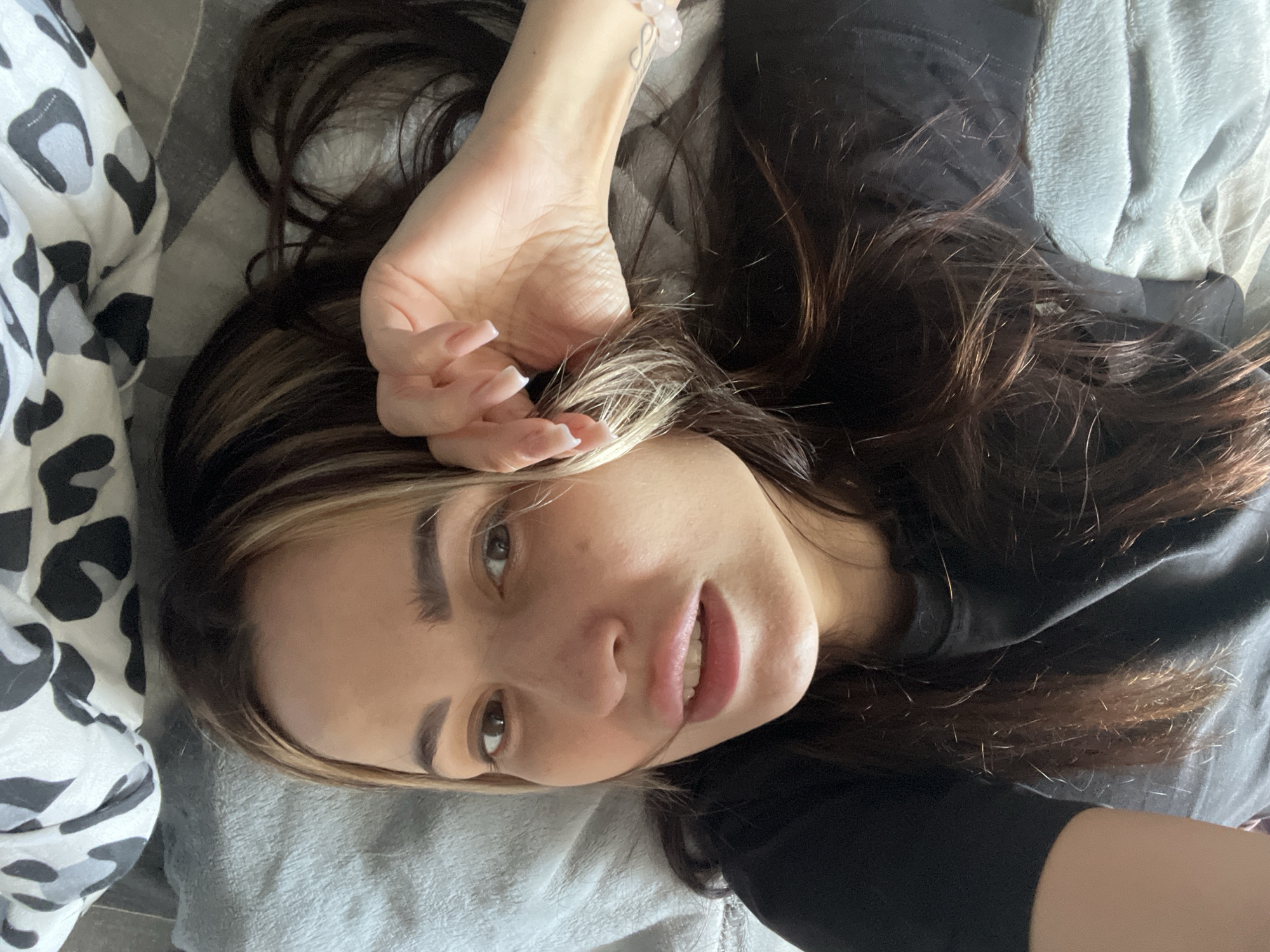 A young woman with brown hair and light highlights is lying on a gray blanket with her head tilted to the left. She is wearing a black shirt and has her right hand resting on her forehead, fingers pointed up. She is looking directly at the camera with a slight smile. To the left of the blanket is a black and white leopard print object. The location is Unknown, Unknown.
