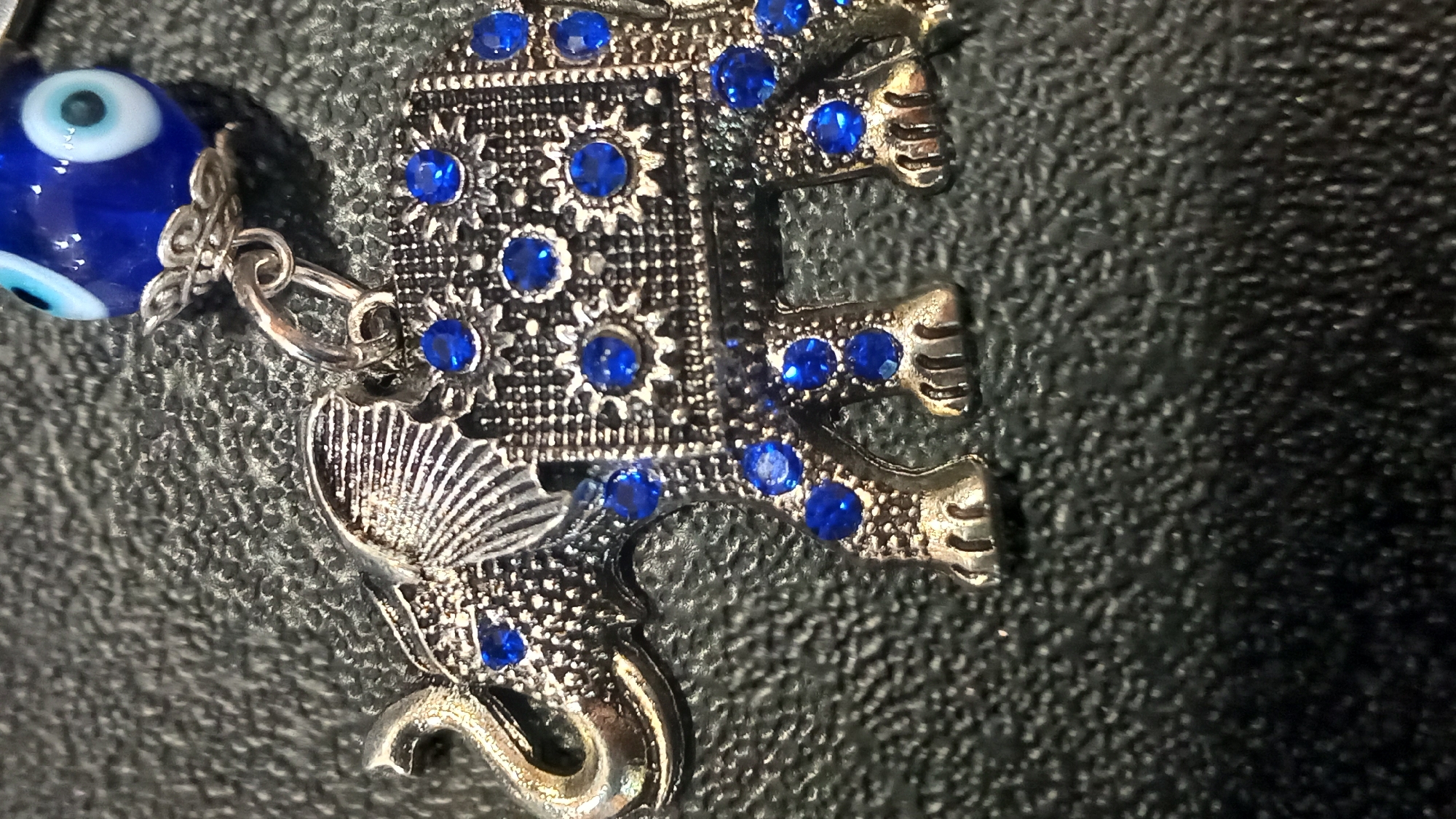 The image shows a close-up of a silver elephant charm adorned with small blue gemstones. The charm is placed on a dark, textured surface, possibly leather or fabric. In the upper right corner of the image, a blue bead with a white and black eye design is partially visible, attached to the elephant charm by a silver chain and ring. The charm is a decorative item, and its exact location within the city of Kos, Greece cannot be determined from the image.