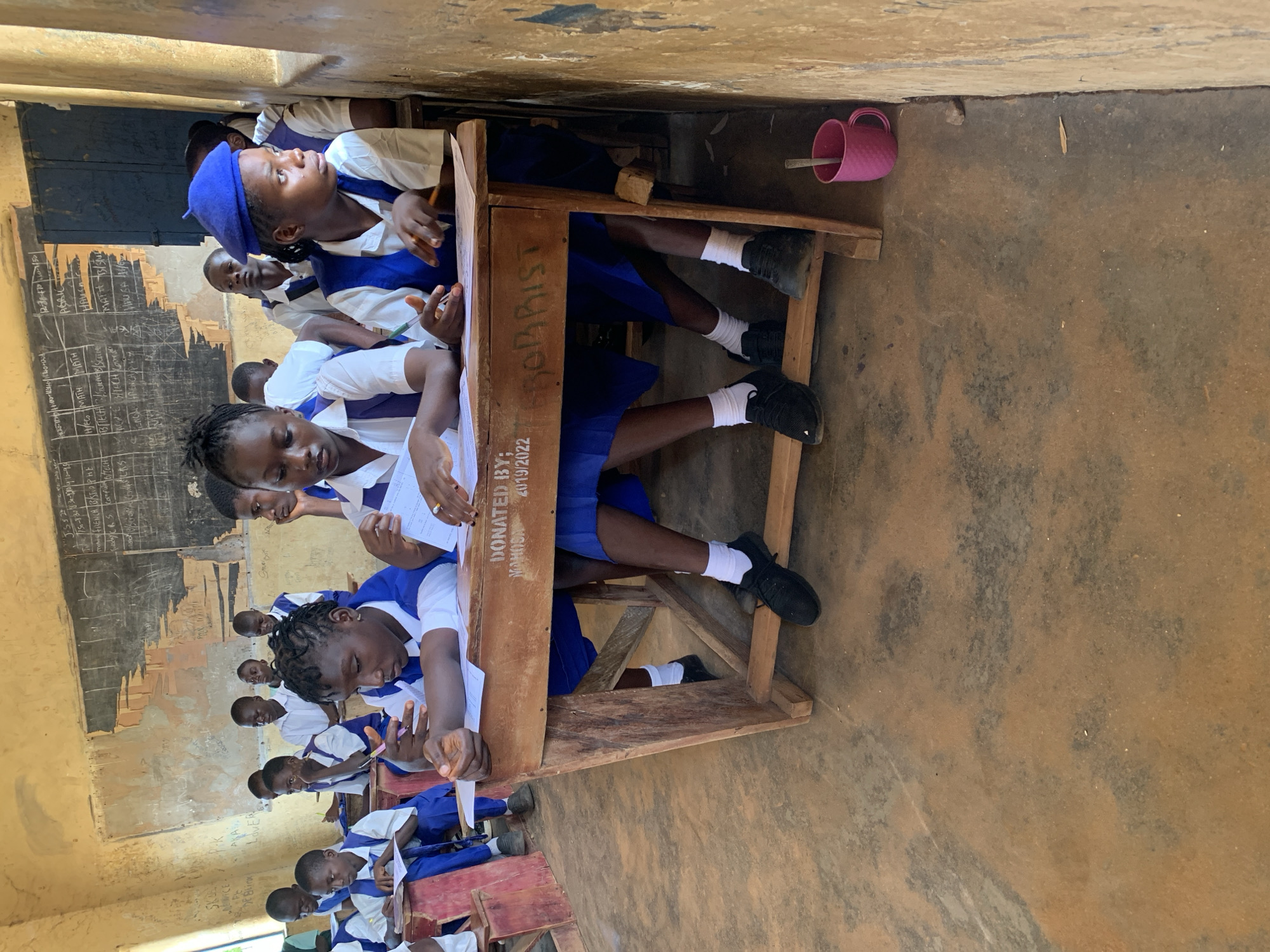 In a classroom in Akwanga, Nigeria, several young students are seated at wooden desks, dressed in blue and white uniforms. The room has a worn chalkboard with writing on it and walls marked with graffiti. One student in the foreground, wearing a blue beret, looks towards the right, her hand resting on a paper. Another student, to her left, is writing on a paper. Several other students are visible in the background, also engaged in writing or looking towards the front. A pink mug with a utensil sits on the floor near the desks. The floor is concrete with a dirt-like texture. The scene captures a moment of learning in a rural educational setting.