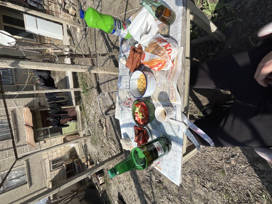 The image captures an outdoor meal setting in a sunlit, unkempt backyard or open area in Kyulevcha, Bulgaria. The scene is viewed from a first-person perspective, with the legs of an individual wearing black pants and dark shoes visible beneath a wooden table.
The wooden table is covered with sheets of newspaper serving as a tablecloth. Arranged on the table are several items: a large green plastic bottle of Kamenitza beer, a smaller green glass bottle, a paper cup containing a dark liquid (likely coffee), and two small patterned glasses, one containing a dark liquid. Food items include a white enamel bowl filled with French fries, a green bowl containing a salad of chopped tomatoes and cucumbers, a white plate with multiple reddish processed meat items (likely sausages), and another white plate with dark red food. A plastic bag from Kaufland contains a round bread item. A small bundle of white plastic cutlery is present, along with a large bright green plastic bottle labeled "DERBY PERO Marty," which appears to be a cleaning product. A small, white, irregular object, possibly garlic, is also on the newspaper.
The background reveals a two-story, light-colored stucco building, presumably a residence, with multiple windows, some featuring exterior awnings or blinds. A ground-floor entrance with a light green door and a brown door is visible under a small canopy. Dark laundry is hanging on a clothesline near the building. The ground is dry, patchy with sparse vegetation, and contains various debris including planks and bricks. Concrete posts with wires are scattered throughout the yard. To the far right, an outdoor white upright refrigerator and a satellite dish are visible near the building.