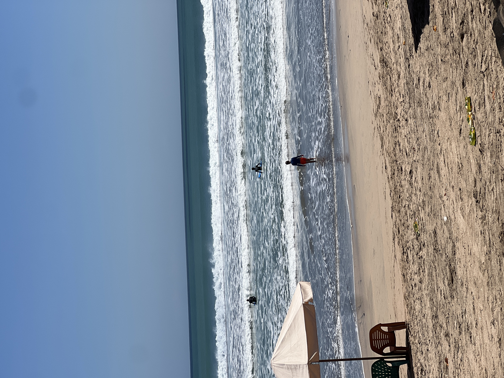 In Kuta, Indonesia, a sunny beach scene unfolds with a bright blue sky above. The ocean stretches out, with multiple rows of waves breaking on the shore. Several surfers are visible in the water, one in the foreground wading into the sea, and others further out with surfboards. To the left, a large tan beach umbrella provides shade over two empty beach chairs, one green and one brown. The sandy beach is mostly bare, with some debris scattered on the ground, including a small offering with yellow flowers.