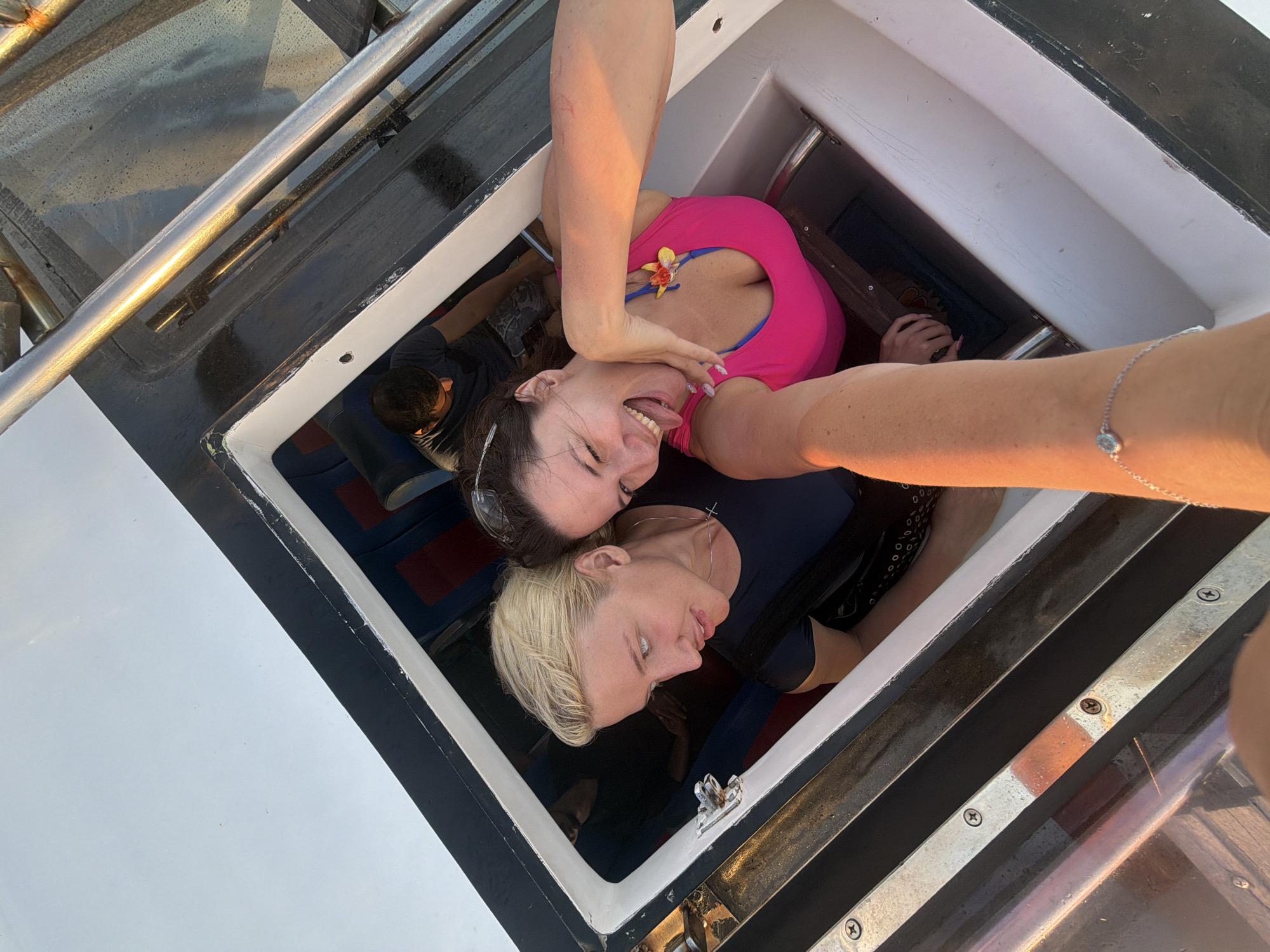 In Denpasar, Indonesia, two women are captured in a high-angle selfie from a boat. The woman in the foreground, on the right, wears a bright pink top adorned with a yellow and blue flower accessory. She is sticking her tongue out playfully, her eyes closed in amusement. Behind her, to the left, another woman with blonde hair and a dark blue t-shirt looks slightly to her left, her expression neutral. In the background, a man in a dark t-shirt is visible, seated on a bench. The scene takes place within the open hatch of a boat, with glimpses of blue and red seating inside. The metallic railings and white hull of the boat are also prominent elements of the environment. The lighting suggests it is either sunrise or sunset, casting a warm glow on the scene.