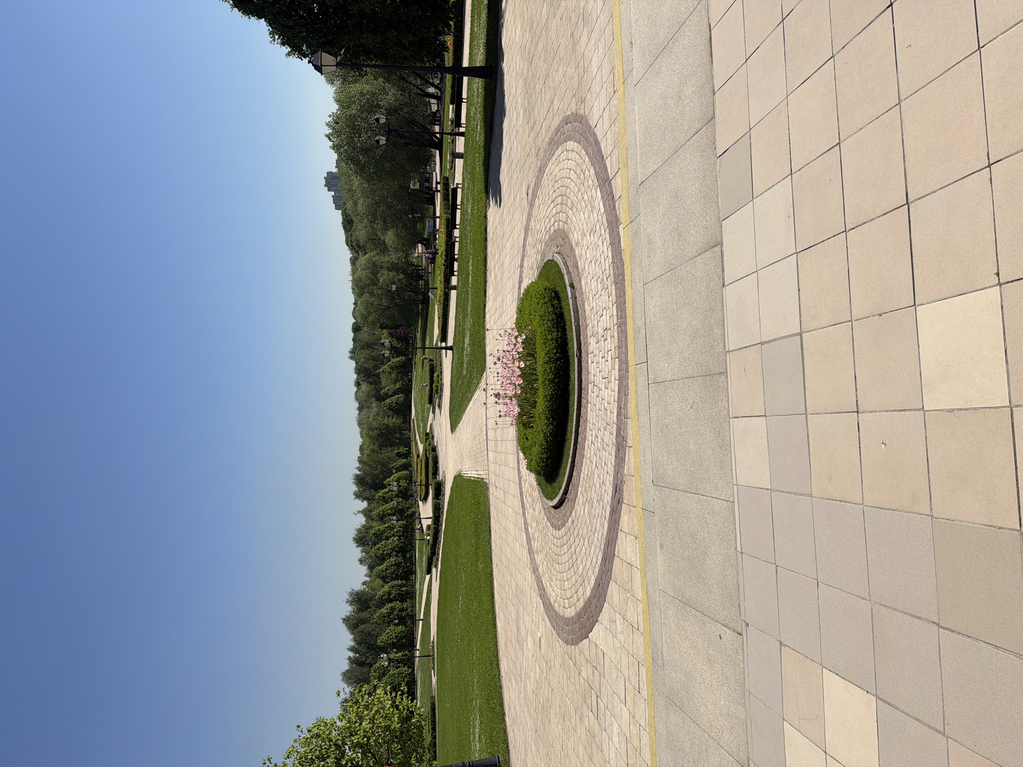 The image depicts a park scene in Moscow, Russia. The camera is positioned at ground level, offering a lengthwise view along a tiled walkway that is offset at an angle. The walkway features a circular arrangement of tiles in its center, enclosing a planting bed with green foliage and pink flowers. Adjacent to the walkway is a strip of grass, flanked by a line of trees and park benches. Lampposts are visible along the path, adding to the park's ambiance. A bright blue sky serves as the backdrop for the scene. The tiled path is made of light-colored, square tiles, while the outer edge of the walkway is lined with a yellow strip. No people are visible in the image.