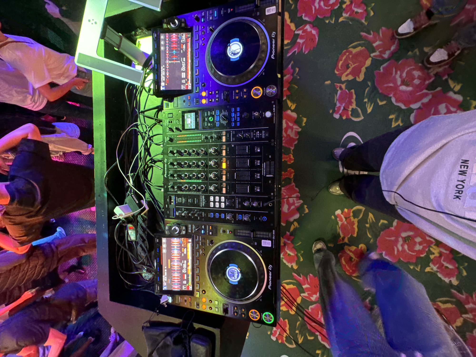 In this overhead shot, a DJ setup is the central focus, positioned on a table with a vibrant floral-patterned cloth. The setup includes two Pioneer DJ CDJ-3000 multi-players and a Pioneer DJM-A9 mixer, all connected by a tangle of black cables. The multi-players display waveforms and track information on their screens. To the left, a black DJ bag rests on the table.
In the background, several individuals are visible, their lower bodies and legs casually dressed, suggesting an informal gathering or event. The lighting appears to be a mix of ambient and stage lighting, with colorful hues like purple and green illuminating the scene. The overall environment seems to be a club or a similar entertainment venue. There is no information that would indicate this location is in Moscow, Russia.