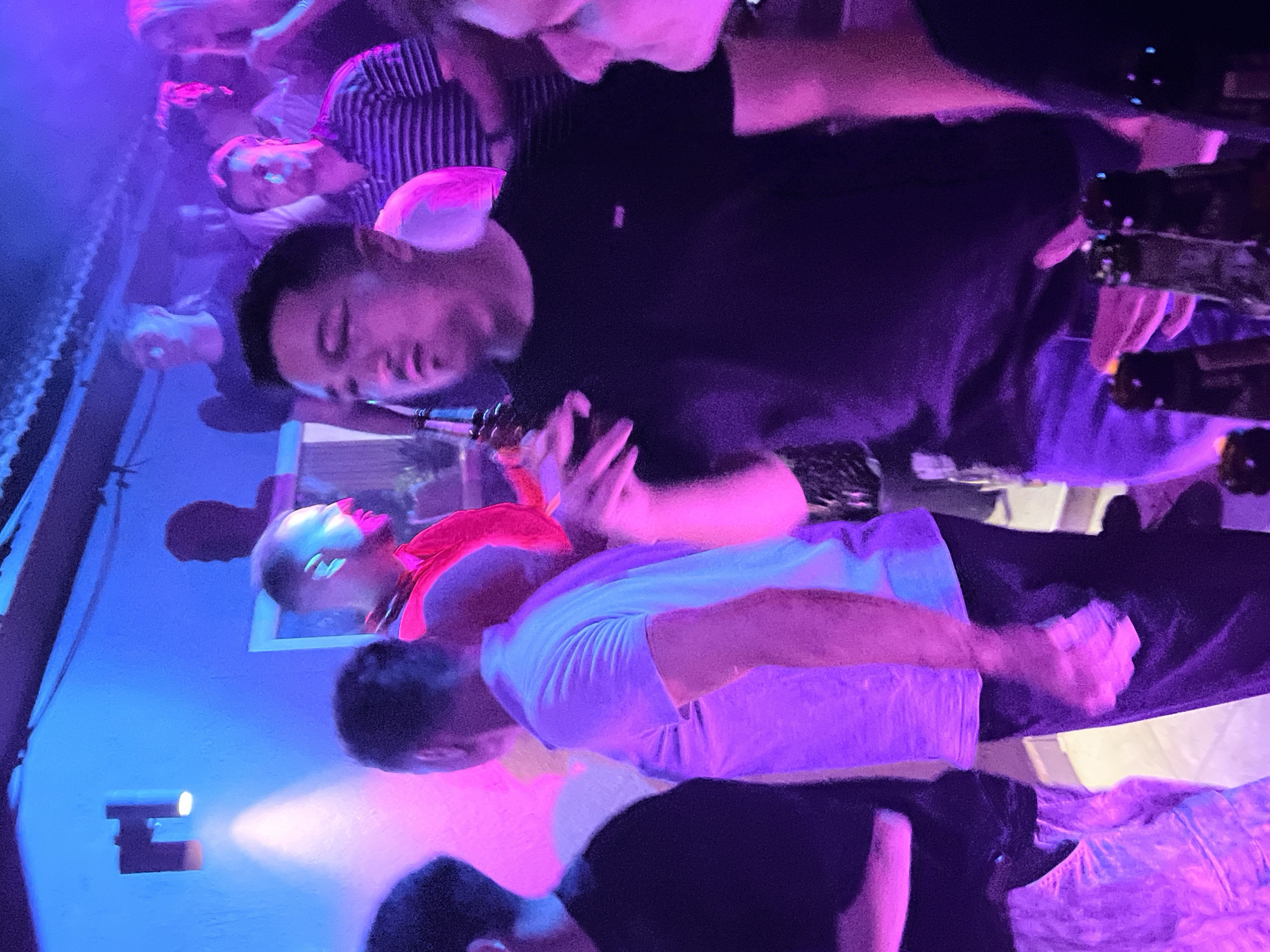The image depicts a dimly lit indoor scene, likely a bar or club, bathed in vibrant blue and pink neon lights. Several individuals are present, with a focus on a man in the foreground wearing a black t-shirt. He is looking away from the camera, with a blurred hand resting on his chest. To his right, another person's face is partially visible, also illuminated by the colored lights. In the background, a group of people are gathered, their features indistinct due to the low lighting and distance. A wall with a light fixture and what appears to be a framed artwork or window is visible on the left. Several bottles, possibly of alcohol, are in the foreground, suggesting a social gathering or event. The overall atmosphere is lively and suggests a nightlife setting. The image does not provide any explicit visual cues to confirm its location in Herceg Novi, Montenegro.