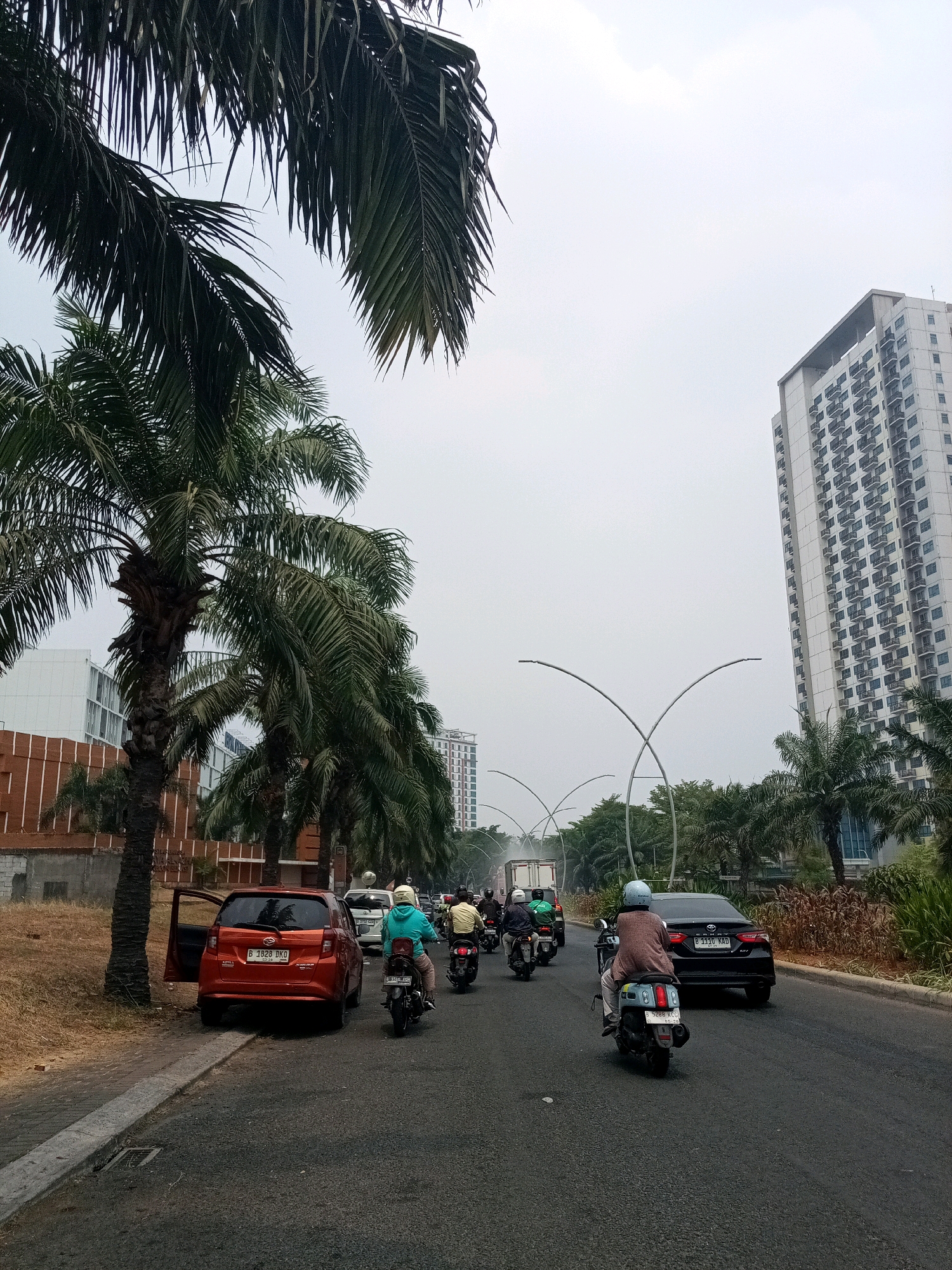 The image depicts a street scene in Bekasi, Indonesia, likely during the daytime under an overcast sky. On the right side of the road, a black sedan with license plate "B 1110 KAD" is in motion. To the left of the sedan, a person is riding a scooter with a light blue helmet and a brown cardigan. Further ahead, several other motorcycles are visible, with riders wearing helmets and casual clothing. One motorcycle has a white license plate that appears to read "B 3533...".
On the left side of the street, a red car with its driver's side door open is parked on the shoulder. Its license plate is "B 1828 DKO". A row of palm trees lines the left side of the road, with some overhanging the sidewalk. In the background, a tall, modern, white apartment building with numerous balconies is visible. The street is paved and appears to be a multi-lane road. Other vehicles, including a white truck, are present in the distance. The overall impression is that of a busy urban environment.
