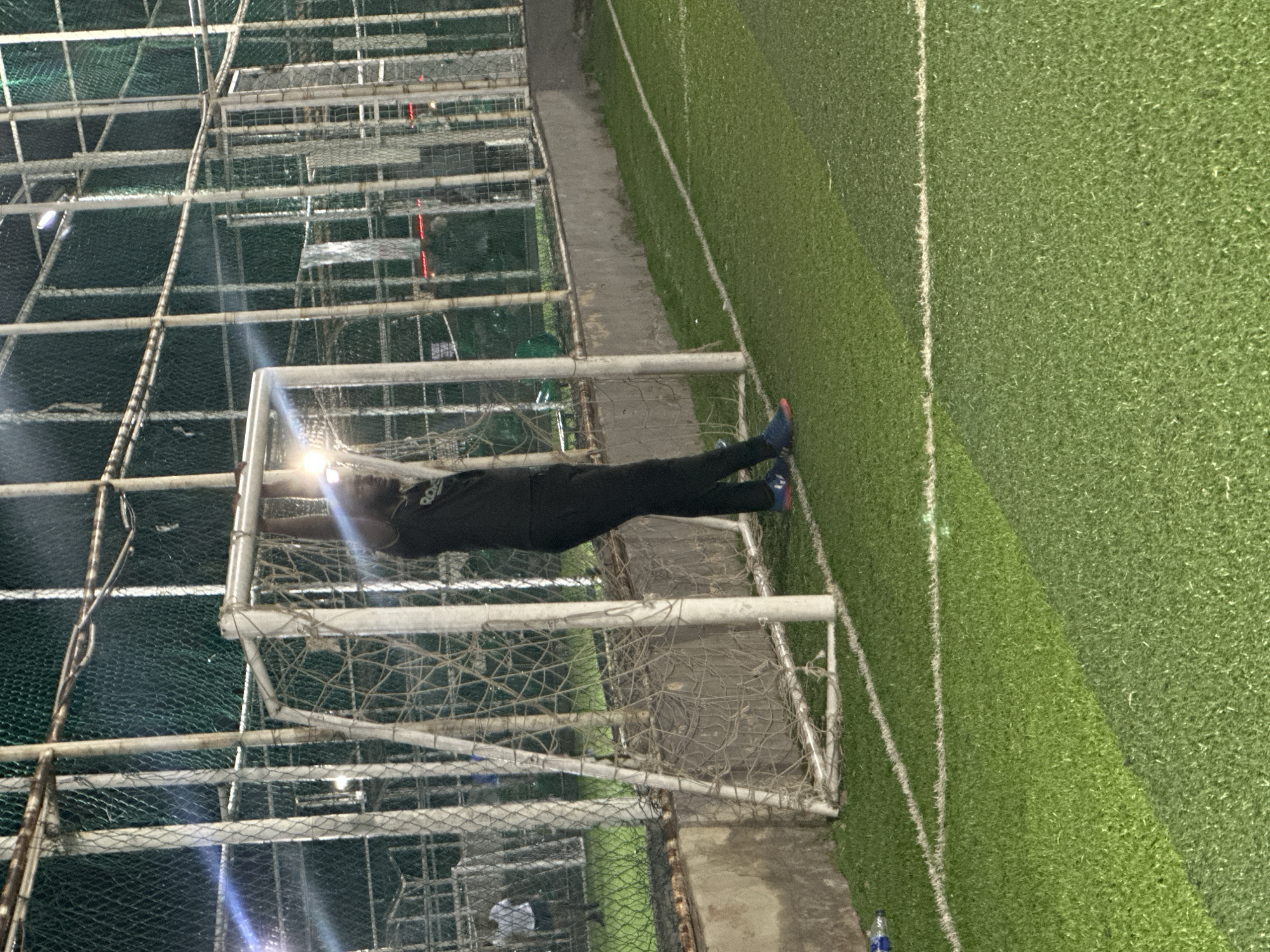 A Black man, wearing a black t-shirt and black pants, stands with his hands on the crossbar of a soccer goal on a green turf field at night. He is positioned to the right of the goal frame. The field is illuminated by overhead lights, creating lens flares and reflections. In the background, a chain-link fence encloses the field, and other structures and lights are visible. Further in the background, a person in a white t-shirt and dark shorts walks on the turf field. The scene is located at a sports facility in Abuja, Nigeria.