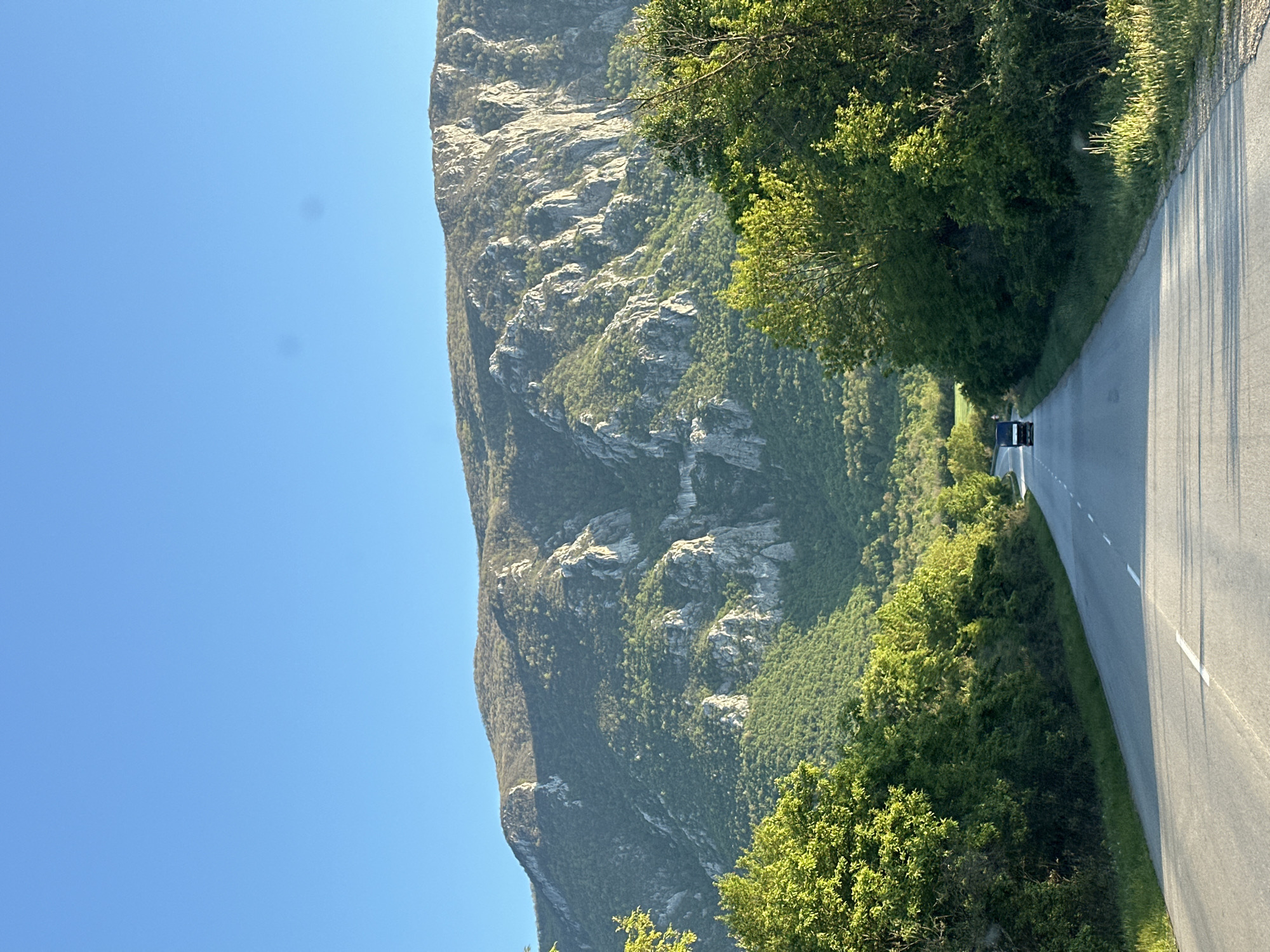 The image shows a road in Kravoder, Bulgaria. The road is asphalt and has white lines. A dark-colored vehicle is driving on the road. To the left of the road is a large mountain with exposed rock faces and green vegetation. To the right of the road are trees and bushes. The sky is clear and blue.