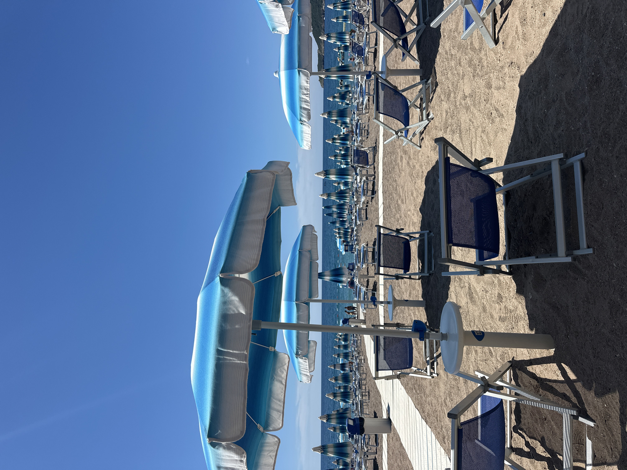 The image depicts a beach scene in Boreti, Montenegro, characterized by rows of blue and white striped beach umbrellas and matching lounge chairs set up on a sandy shore. In the foreground, several lounge chairs are positioned on the sand, some with small white tables attached to their central poles. A white wooden walkway extends into the background. The sea is visible beyond the chairs and umbrellas, with a clear blue sky above. In the middle ground, a few people are visible near the water's edge. The overall impression is one of a well-maintained and prepared beach ready for visitors.