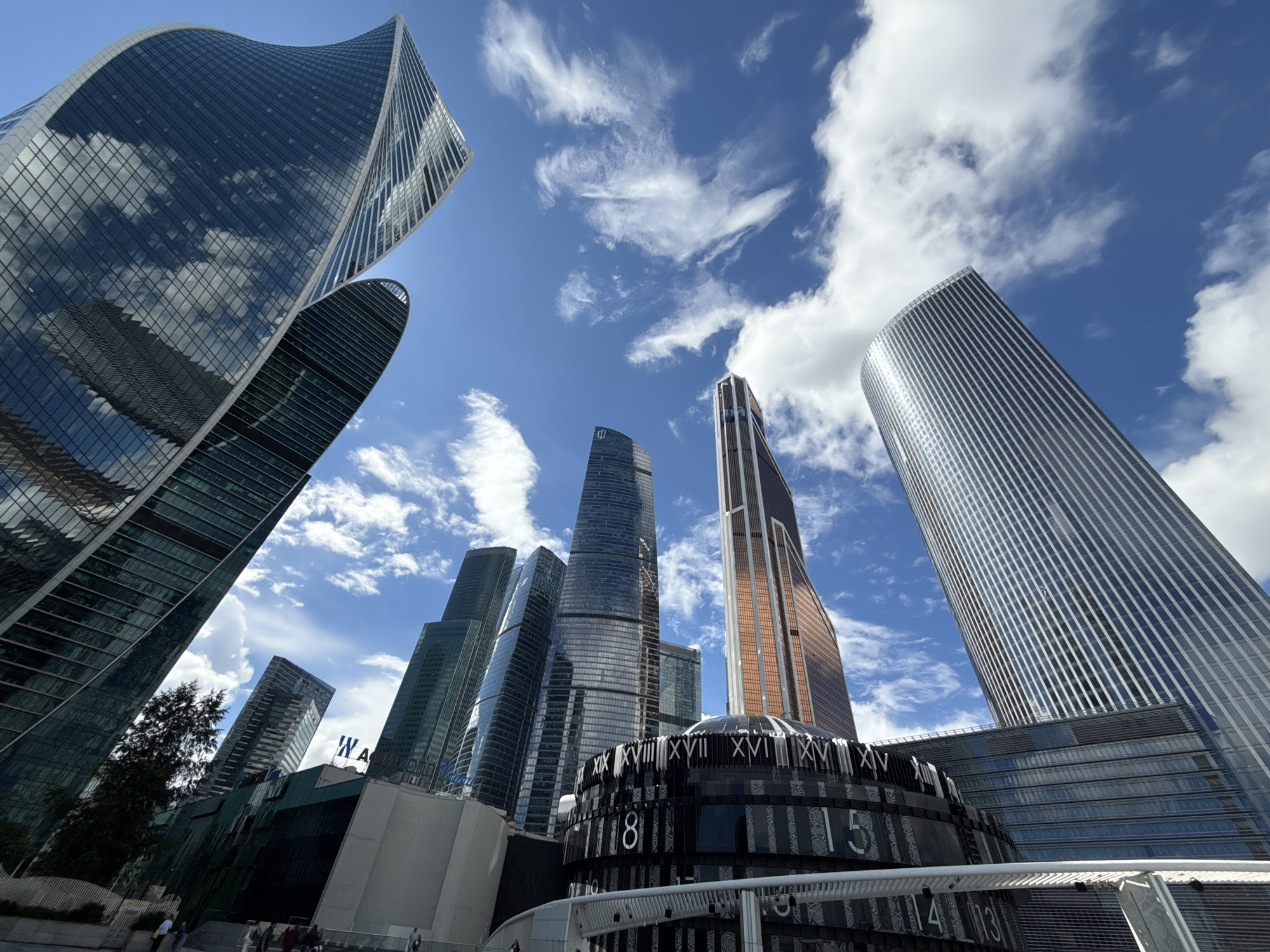 This image captures a low-angle perspective of the Moscow International Business Center, also known as Moscow-City, in Moscow, Russia. The foreground features a distinctive cylindrical structure with a clock face displaying Roman numerals. Several modern skyscrapers, characterized by their glass facades and varying architectural designs, dominate the background. The sky is a vibrant blue with scattered white clouds, suggesting a clear day. The buildings reflect the sky and clouds, adding depth and visual interest to the scene. A few individuals are visible walking in the lower part of the image, indicating a public urban space. The overall scene is one of a contemporary architectural hub within a bustling city.