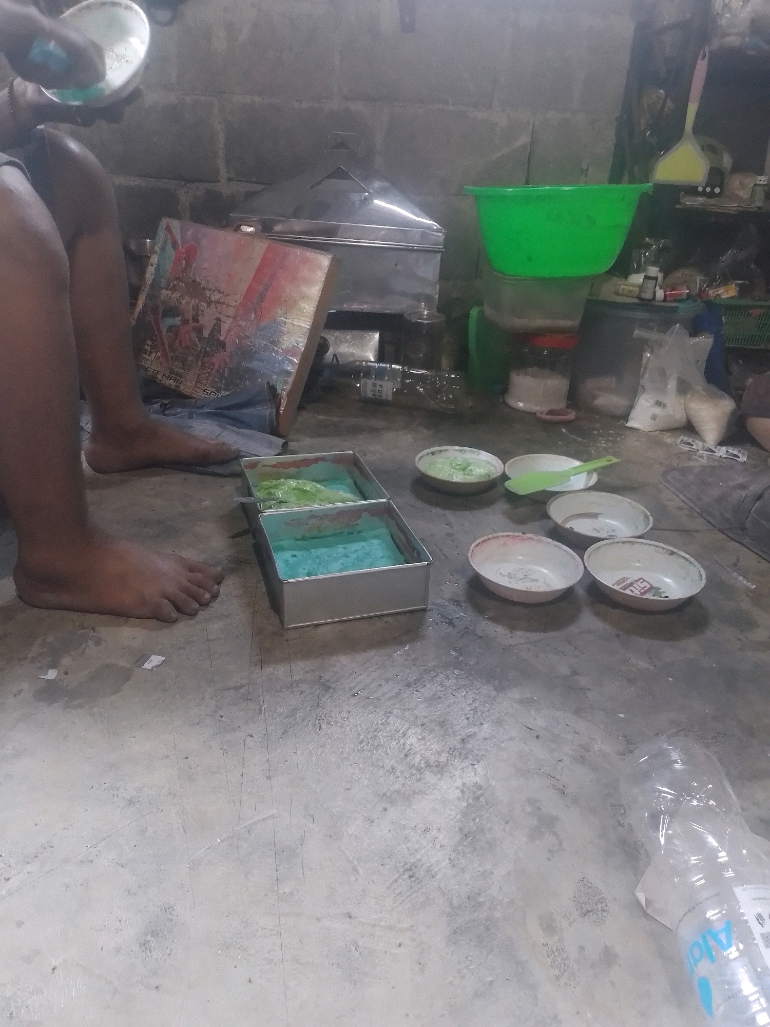 In an indoor setting with a concrete floor and a brick wall, a person's legs are visible, positioned to the left of the frame. The person is barefoot. In front of the legs, there is a rectangular metal pan containing two compartments filled with a blue and green substance. Several small, shallow bowls are scattered on the floor to the right of the pan. One bowl contains a green substance, and another contains a light green substance. A green spatula is placed across two of the bowls. Further to the right, a large green plastic bowl is stacked on top of a clear plastic container. Behind these items, there are shelves with various objects, including bags of what appears to be rice, bottles, and a yellow dustpan with a pink handle. A metallic steamer is visible behind the person's legs. To the far right, a clear plastic water bottle lies on its side. A colorful cardboard is leaning against a wall in the background. The scene appears to be a workspace where some kind of food preparation or craft activity is taking place.