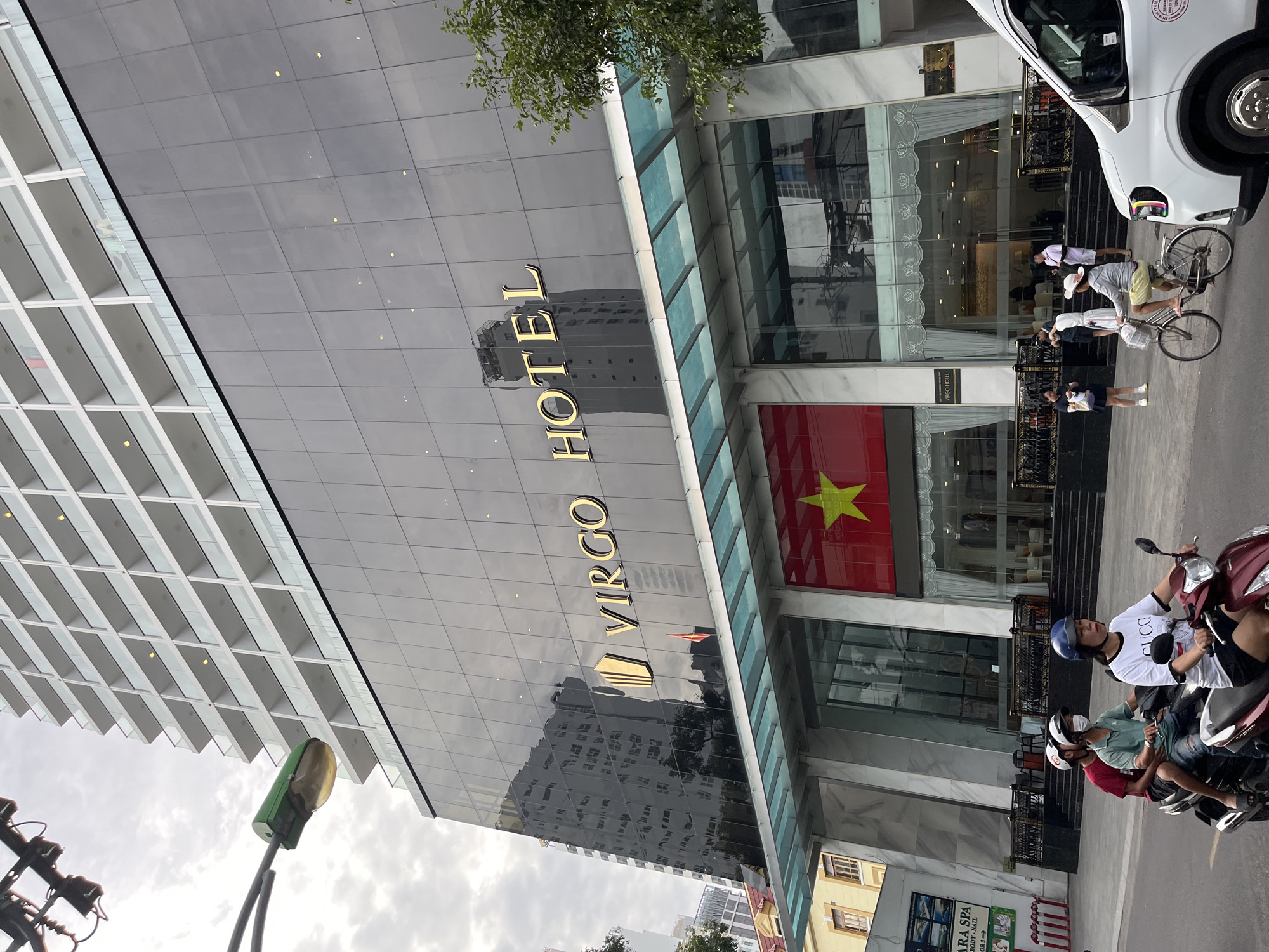 The image shows the exterior of the Virgo Hotel in Nha Trang, Vietnam. The hotel features a modern facade with a glass exterior and gold lettering that reads "VIRGO HOTEL." A Vietnamese flag is displayed prominently on the building. In front of the hotel, people are seen walking and riding motorcycles, reflecting the typical street life of the city. A white van is parked on the side of the road, and a tree with green foliage adds a touch of nature to the urban landscape. The sky is overcast, suggesting a cloudy day.