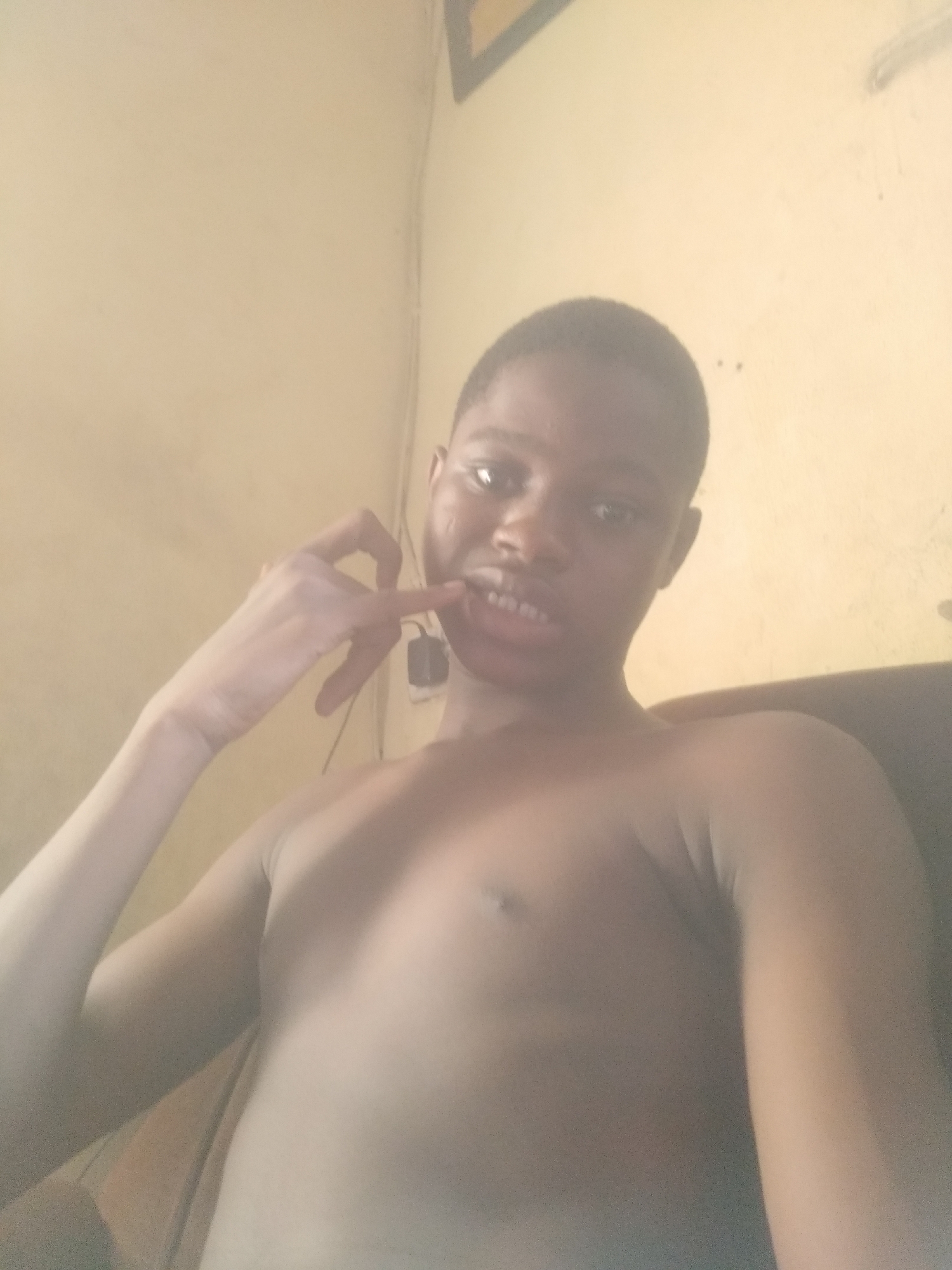 The image features a young individual, likely male, with dark skin and short, dark hair, captured in a close-up, chest-up shot. He is shirtless, revealing his upper torso and arms. The subject is looking directly at the camera with his mouth slightly open, showing his teeth. His left hand is raised, with the index finger pointing towards his mouth, suggesting a playful or casual pose. A small, dark, rectangular object, possibly an earbud, hangs near his left shoulder.
The setting appears to be indoors, against a light-colored, warm-toned wall that shows some faint textures and possibly a dark, angular shape in the upper right corner, which could be part of a frame or furniture. Behind the subject's right shoulder, a dark, possibly upholstered surface is visible, suggesting he is sitting on a chair or bed. The lighting is soft and diffused, creating a slightly warm and hazy atmosphere. No specific time of day or weather conditions can be discerned, and there is no visible text in the image.