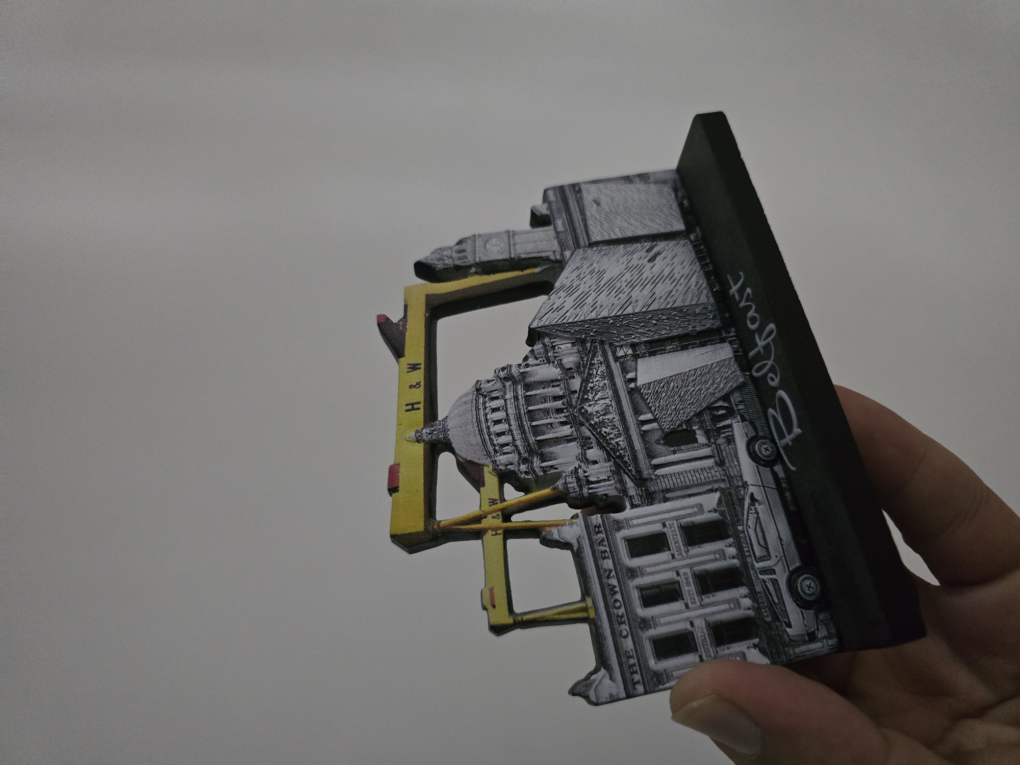 A hand is holding a 3D souvenir depicting landmarks from Belfast, Northern Ireland. The souvenir features the Samson and Goliath cranes, Belfast City Hall, and the Crown Bar. The words "Belfast" are written in white script on the base of the souvenir. The background is a plain, light gray wall. The souvenir is illuminated from the left, casting a subtle shadow to the right. There is no visible connection to Qaratau, Kazakhstan.