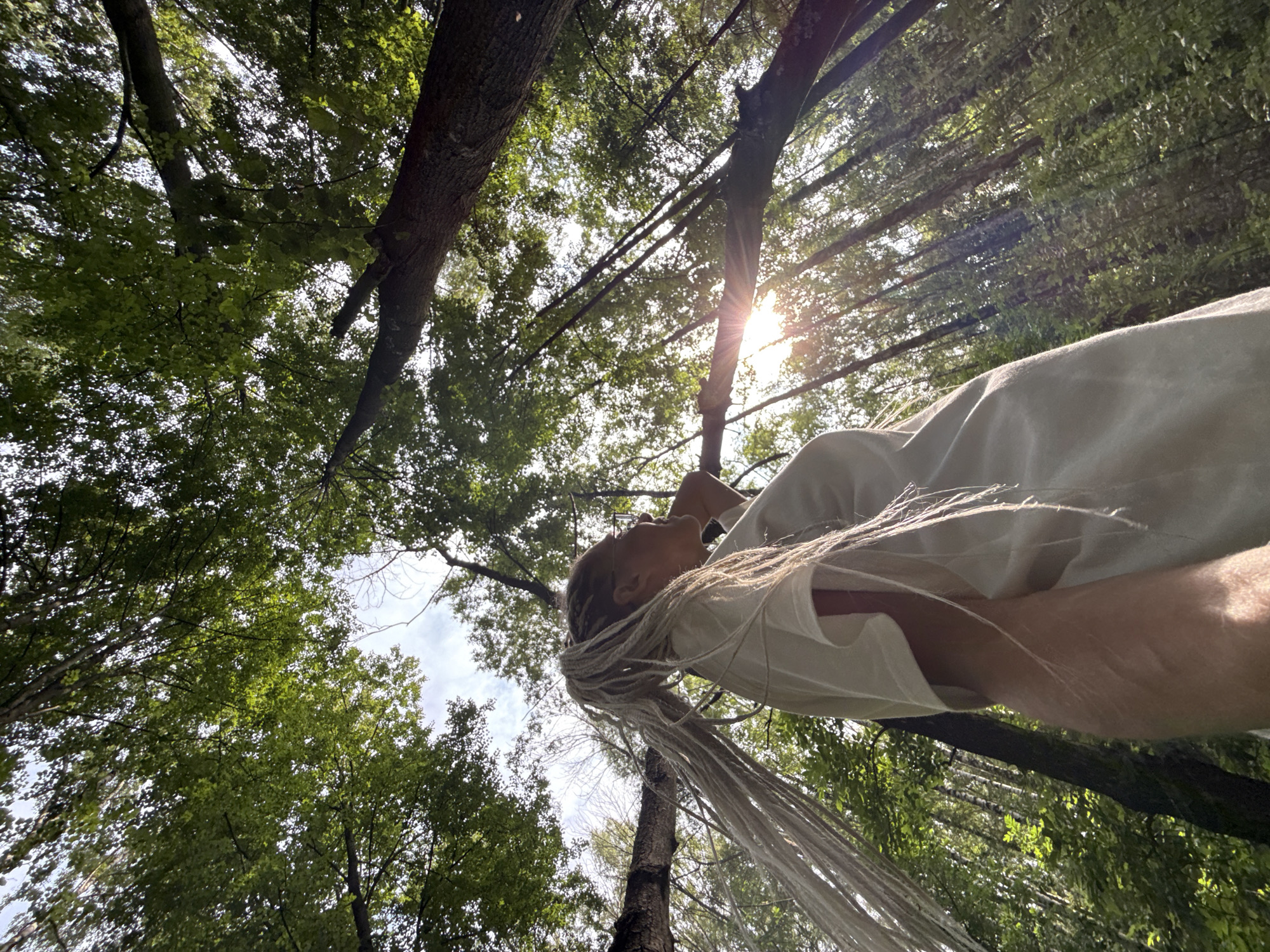 A low-angle shot captures a woman with blonde dreadlocks looking upwards through a canopy of green trees. The sun shines through the branches, creating a lens flare effect. The woman is wearing a light-colored t-shirt. The background shows a dense forest with sunlight filtering through the leaves. The scene appears to be in a rural area.
