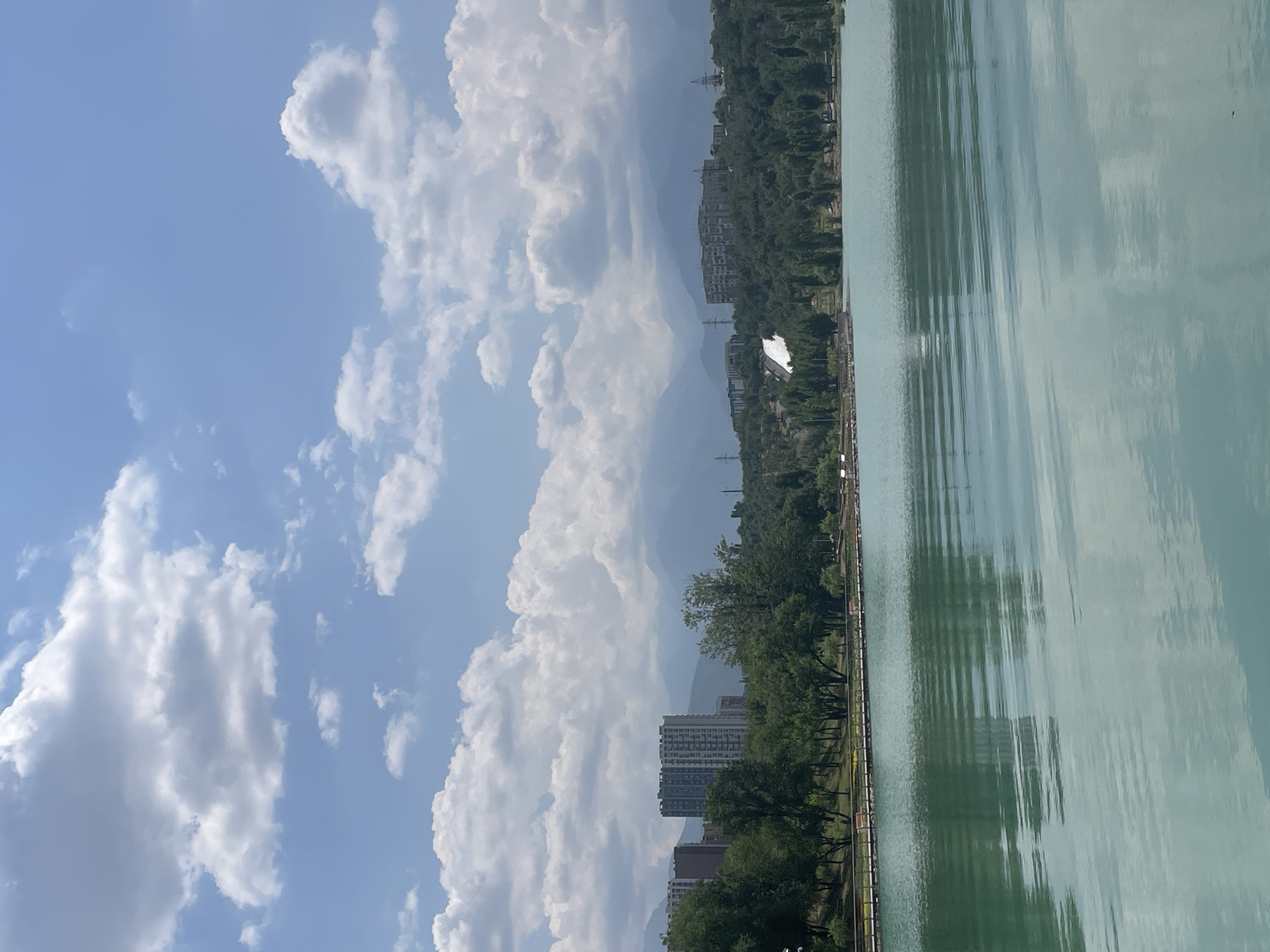 The image depicts a wide, serene body of light turquoise water in the foreground, reflecting the sky and surrounding elements. In the midground, a tree-lined embankment is visible, with a walkway featuring benches and a railing. Behind the embankment, a lush green forest covers rolling hills. Several modern, multi-story buildings rise above the trees, some appearing to be residential complexes, with one particularly prominent, light-colored building with a distinctive facade. In the background, a range of hazy blue mountains stretches across the horizon. The sky is a bright blue, dotted with fluffy white clouds. The scene appears to be a scenic park or recreational area, likely within or on the outskirts of a city. Based on the distinctive mountain range and architectural styles, this location is likely Kok-Tobethe park in Almaty, Kazakhstan, a popular urban park offering panoramic views of the city and the Tien Shan mountains.