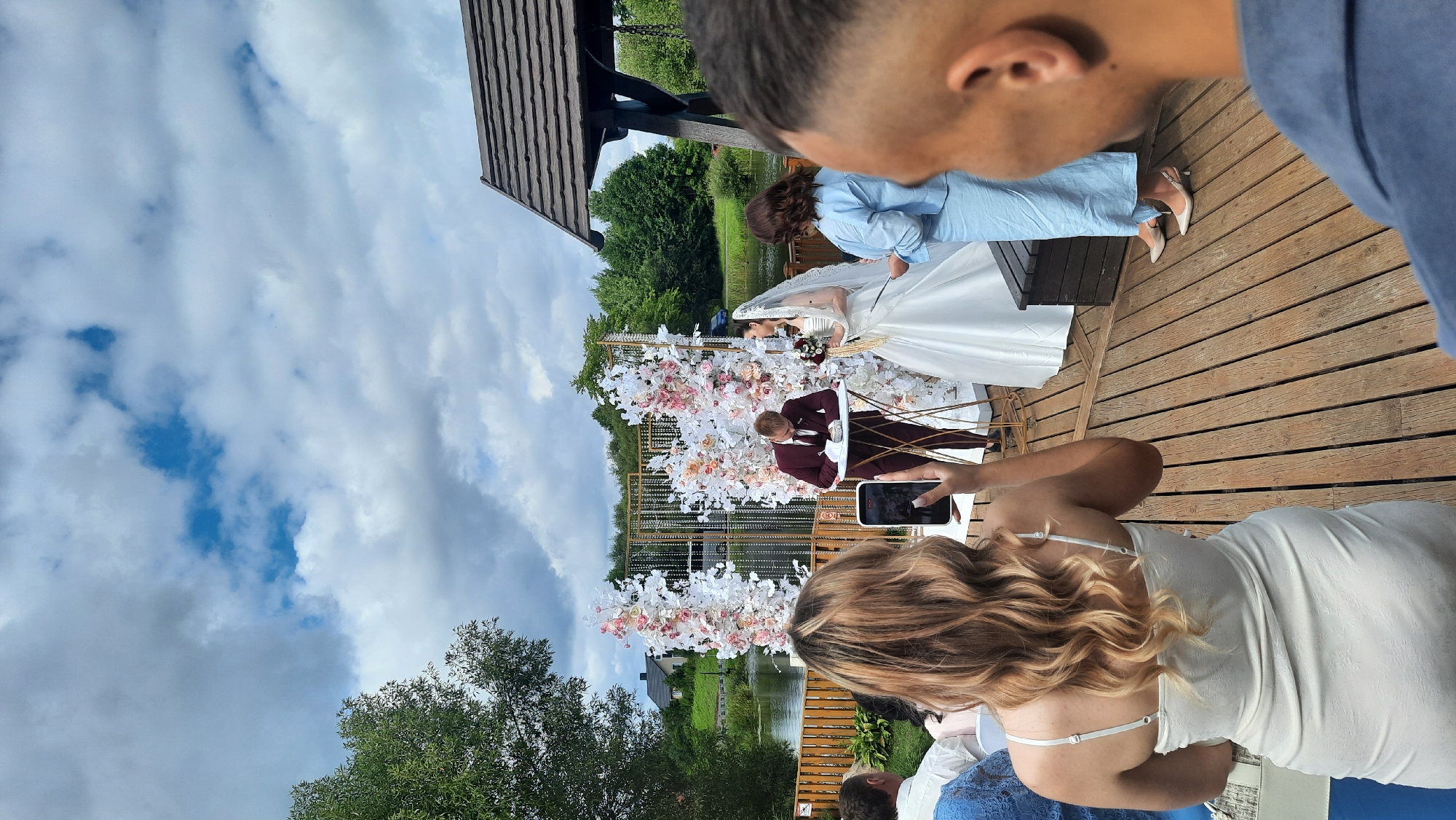 A wedding ceremony is taking place outdoors, on a wooden deck overlooking a body of water, possibly a lake, in the vicinity of Жодзішкаўскі сельскі Савет, Belarus. A bride in a white gown and veil stands to the right of a man in a burgundy suit who is signing a document at a round table. They are positioned in front of a decorative arch adorned with white and pink flowers. Several guests are seated and standing, observing the ceremony. In the foreground, a woman in a light-colored dress is holding up a smartphone, capturing the event. The sky is cloudy with patches of blue. Trees and a building are visible in the background.