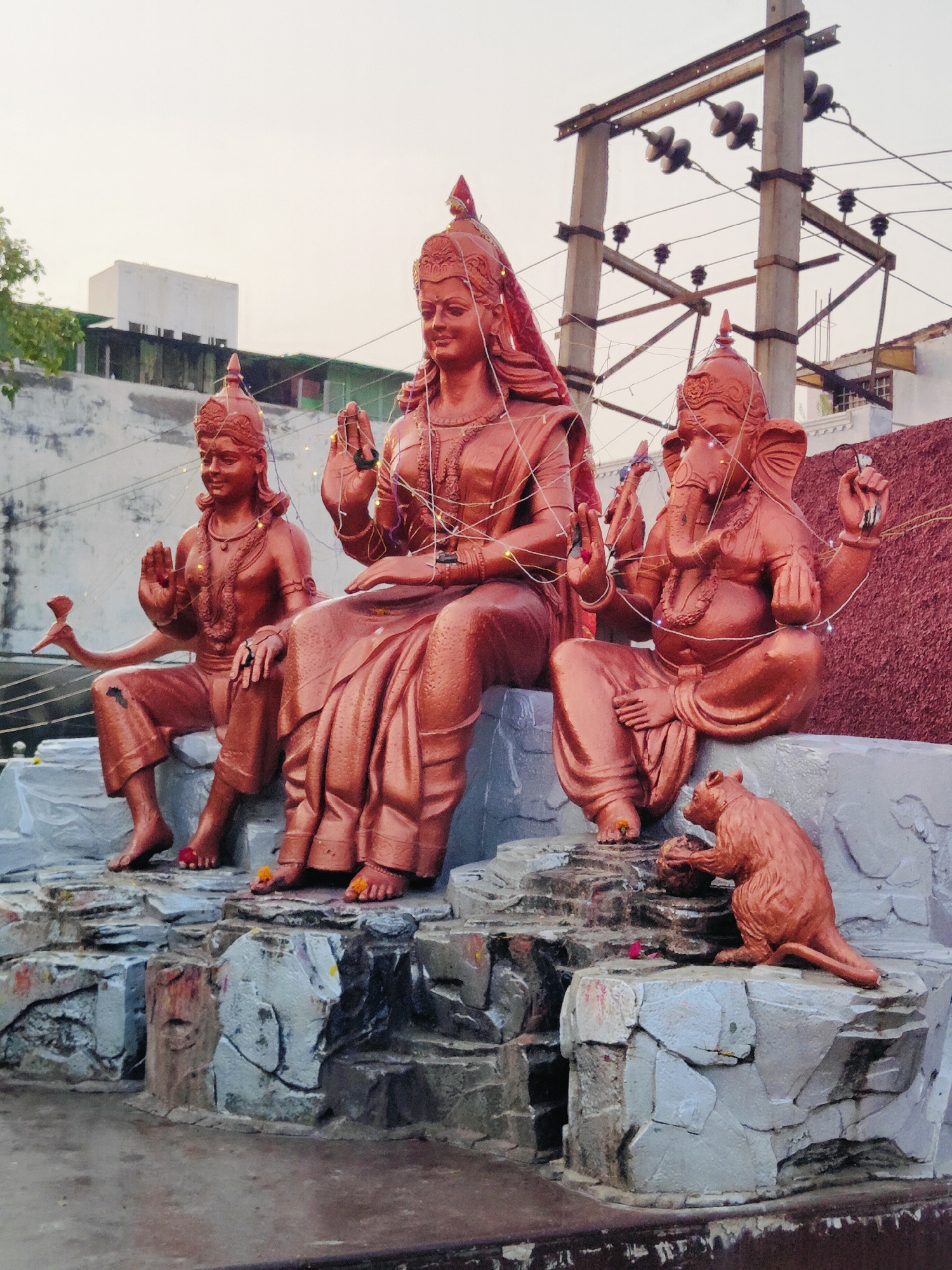 This image captures a religious sculpture group in Gurgaon, India. The central figures are deities from Hinduism, depicted in bronze-toned material. On the left is a male figure with a serene expression, wearing a crown and ornate jewelry, holding his hand up in a blessing gesture. To his right sits a larger female figure, also adorned with a crown and jewelry, with a gentle smile and hands raised in a blessing posture. She is draped in a flowing sari. To the right of the female figure is Ganesha, the elephant-headed god, seated with his trunk and tusks visible, holding an object in his hand and also in a blessing pose. In front of Ganesha, a small creature resembling a rat or mouse is depicted in a crouching position, interacting with a round object. The sculptures are seated on a tiered base made of grey, rock-like material, with some red decorative elements and small floral offerings. The background shows a light-colored building with windows, electrical poles with wires, and a hazy sky, suggesting an outdoor urban setting. The overall scene is illuminated by natural light, possibly during dusk or dawn.