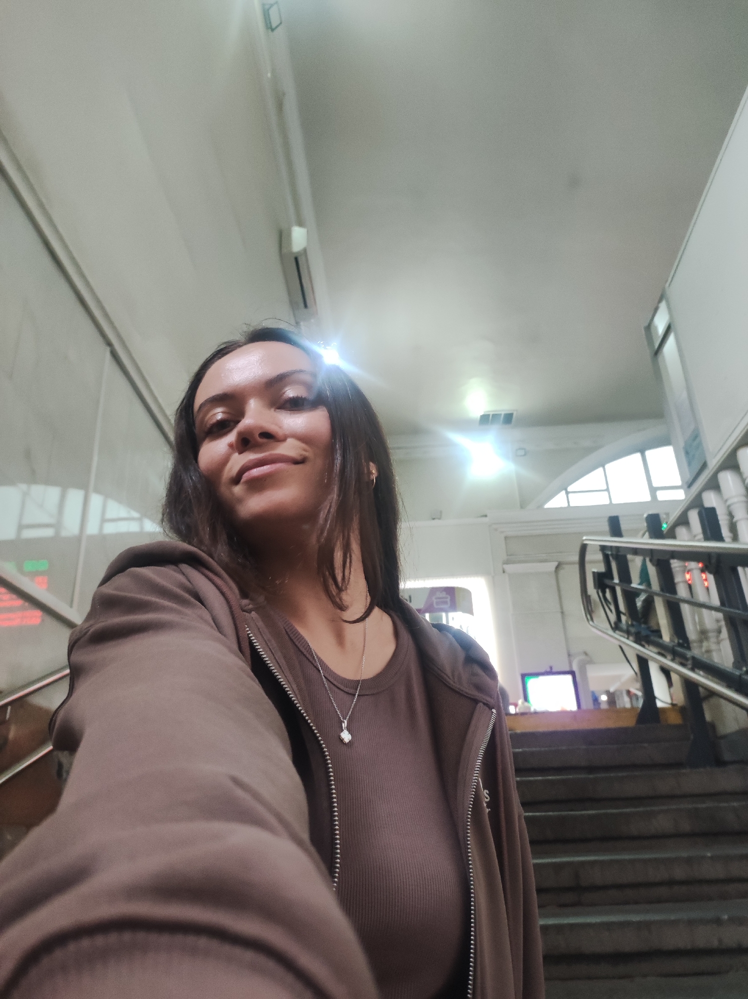 A young woman with long, dark hair is taking a selfie in a public indoor space, likely a transit station or building with stairs. She is wearing a brown zip-up hoodie over a brown ribbed top, and a silver necklace with a small white pendant. The background shows a staircase, handrails, windows with arched frames, and fluorescent lighting on the ceiling. There are also signs with Cyrillic text visible. The lighting is bright, with some glare reflecting off surfaces. The woman is smiling and looking at the camera.