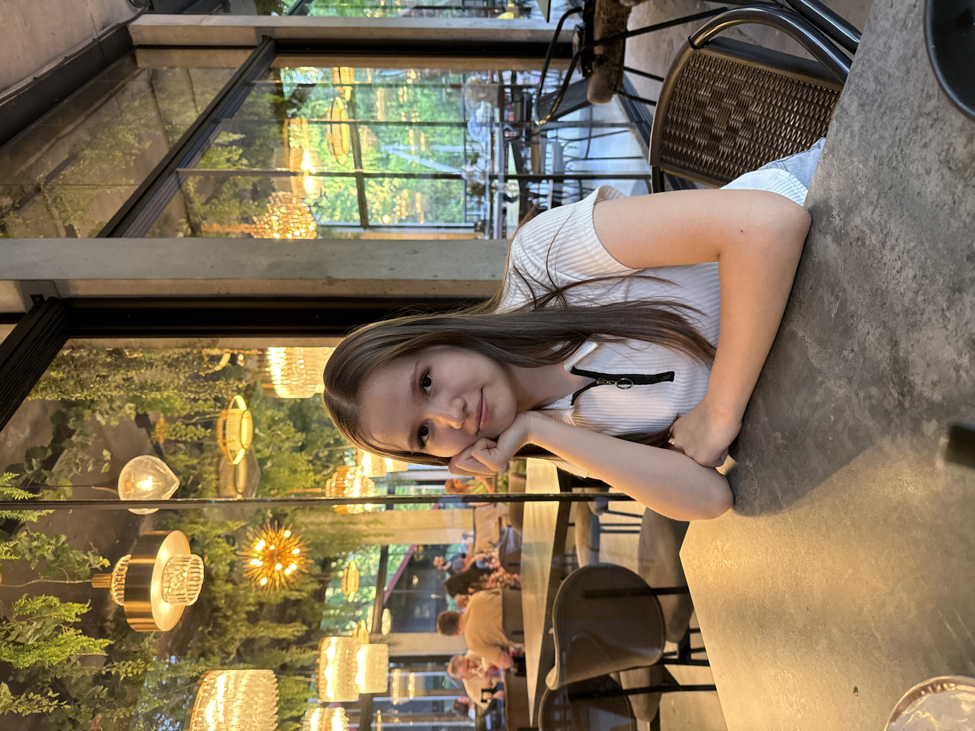 A young woman is seated at a table in what appears to be a restaurant. She is resting her head on her hand, smiling, and looking directly at the camera. The restaurant features a wall of windows showcasing greenery and decorative light fixtures. Other patrons and chairs are visible in the background. The location is unknown, and the city of Izhevsk, Russia, cannot be determined from the given image.