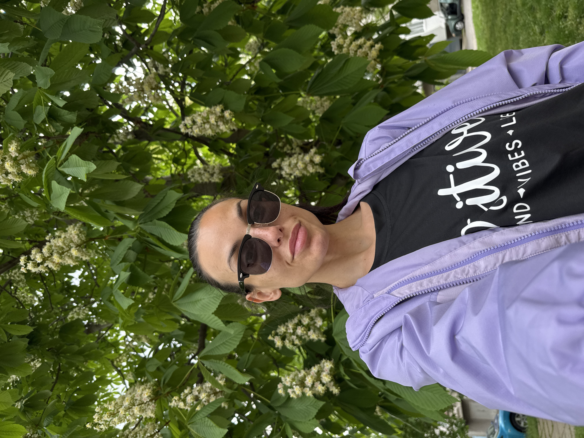A woman is photographed outdoors in Pleven, Bulgaria. She wears sunglasses, a black T-shirt with white lettering, and a lavender jacket. She appears to be standing beneath a tree with green leaves and white flowers. In the background, there is grass, a building, and a partially visible vehicle. The photograph is slightly rotated to the left.