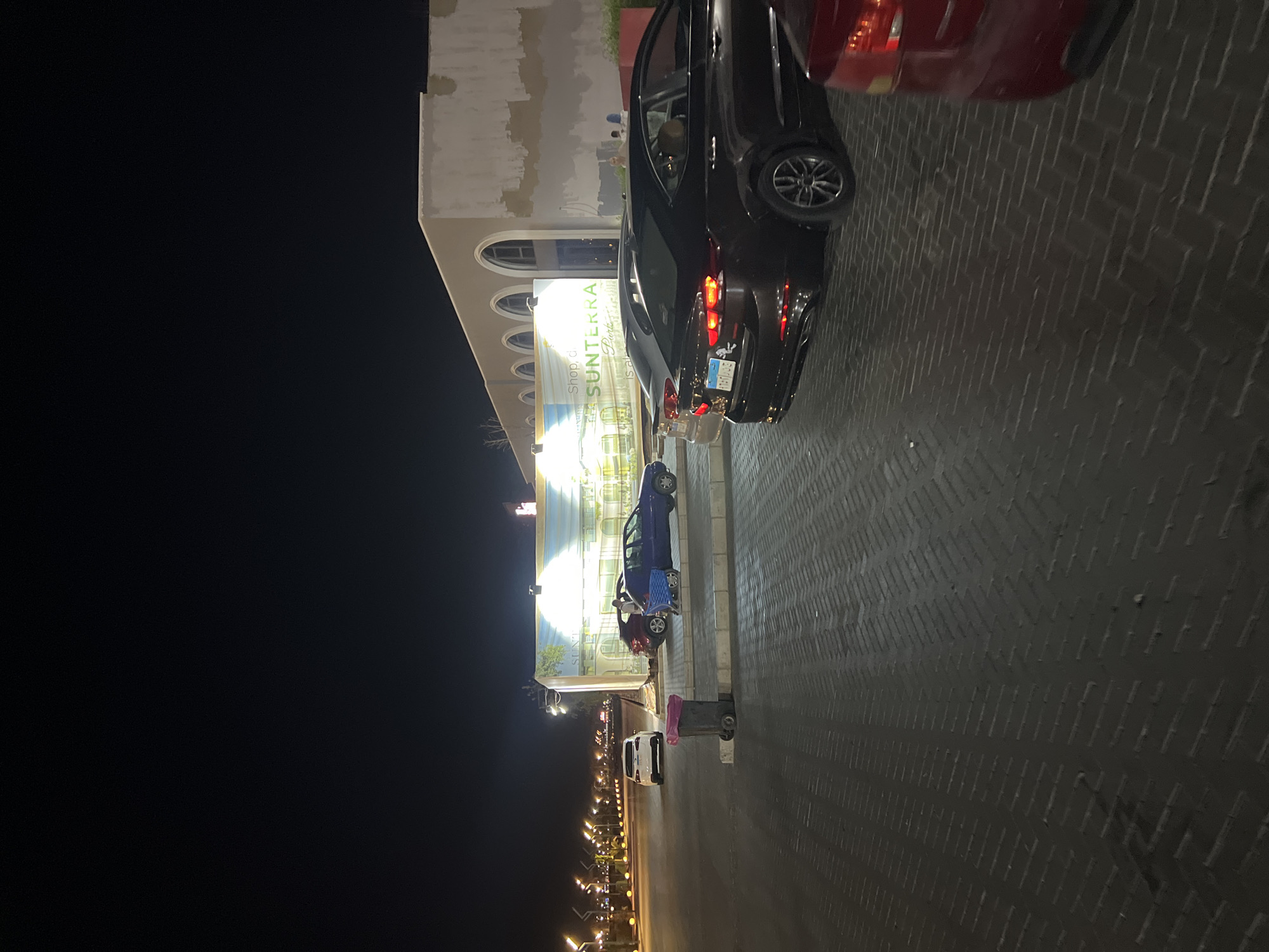 The image shows a nighttime scene in Sharm El Sheikh, Egypt, in front of a building with arched windows. In the foreground, a dark-colored sedan is parked on a patterned brick surface. Further back, a red car is partially visible. To the left, a white Tesla sedan is parked. A blue shopping cart is next to a parked blue sedan, and a red garbage bin is located near the white Tesla. The building behind the vehicles features a mural or large advertisement with text that includes "Shop, dine," and "SUNTERRA Parks." The overall environment appears to be a parking area or plaza.