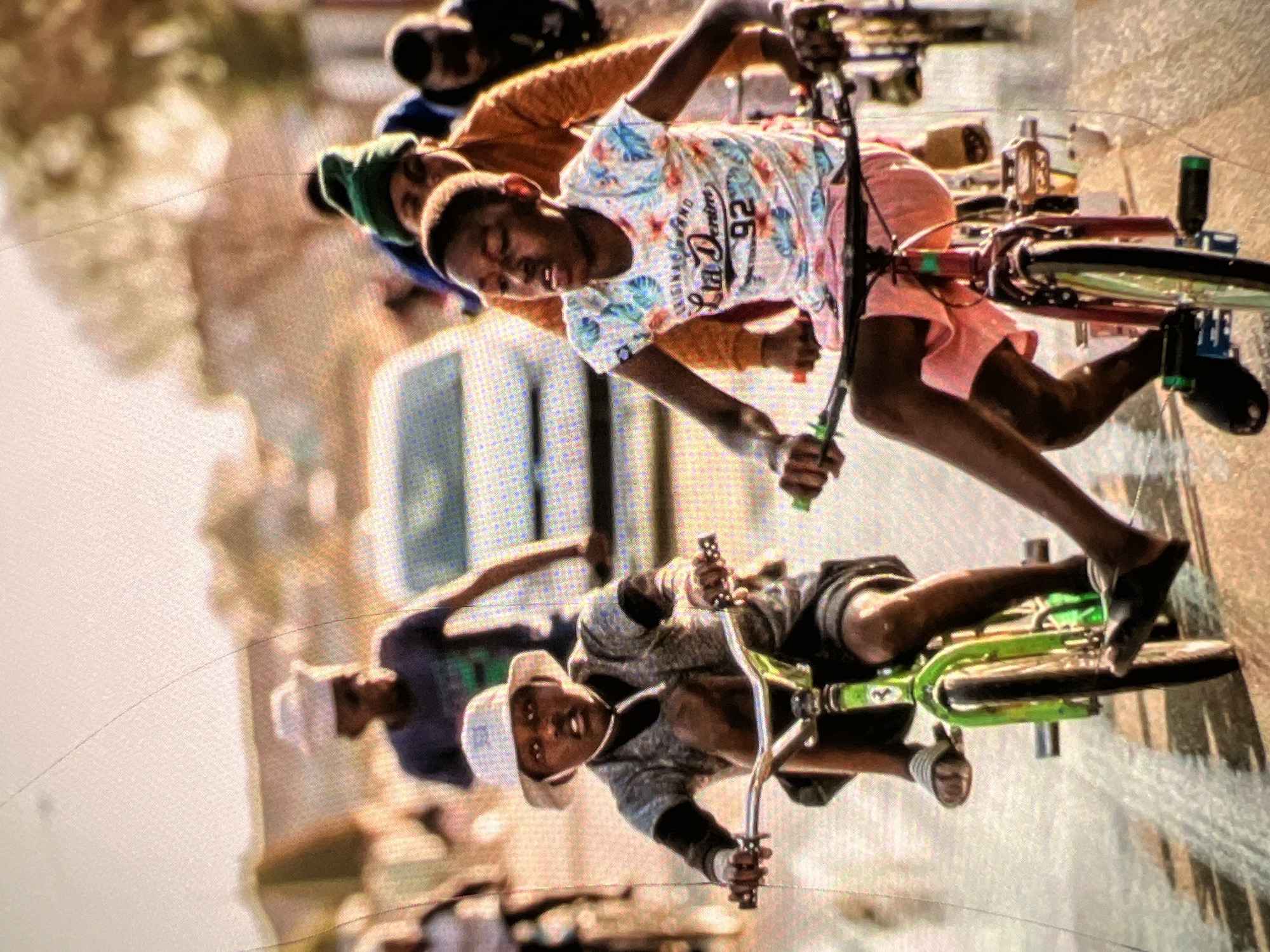 In a city street in Lagos, Nigeria, several young individuals ride bicycles. In the foreground, two boys on bicycles are prominently featured. The boy on the left, wearing a bucket hat, a grey patterned shirt, and dark shorts, is on a green bicycle. The boy on the right, with a green beanie and an orange jacket, is riding a darker colored bicycle and wears a floral t-shirt with pink shorts. Behind them, other children and adults are visible, some also on bicycles, and a car is present in the background. The street surface appears wet, reflecting the light. The overall scene depicts a casual outdoor activity among children in a developing urban environment.